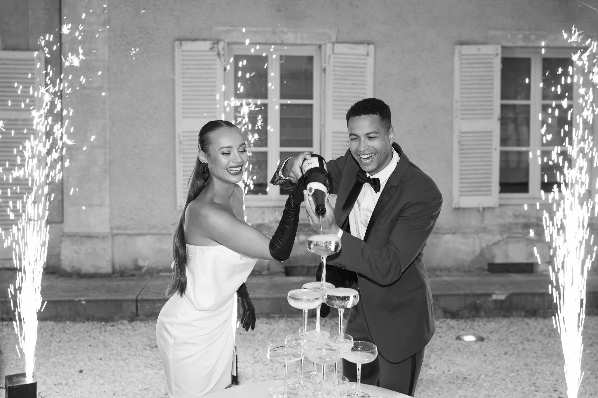 Bride and groom pouring champagne tower at outdoor wedding reception with illuminated wire-frame lights