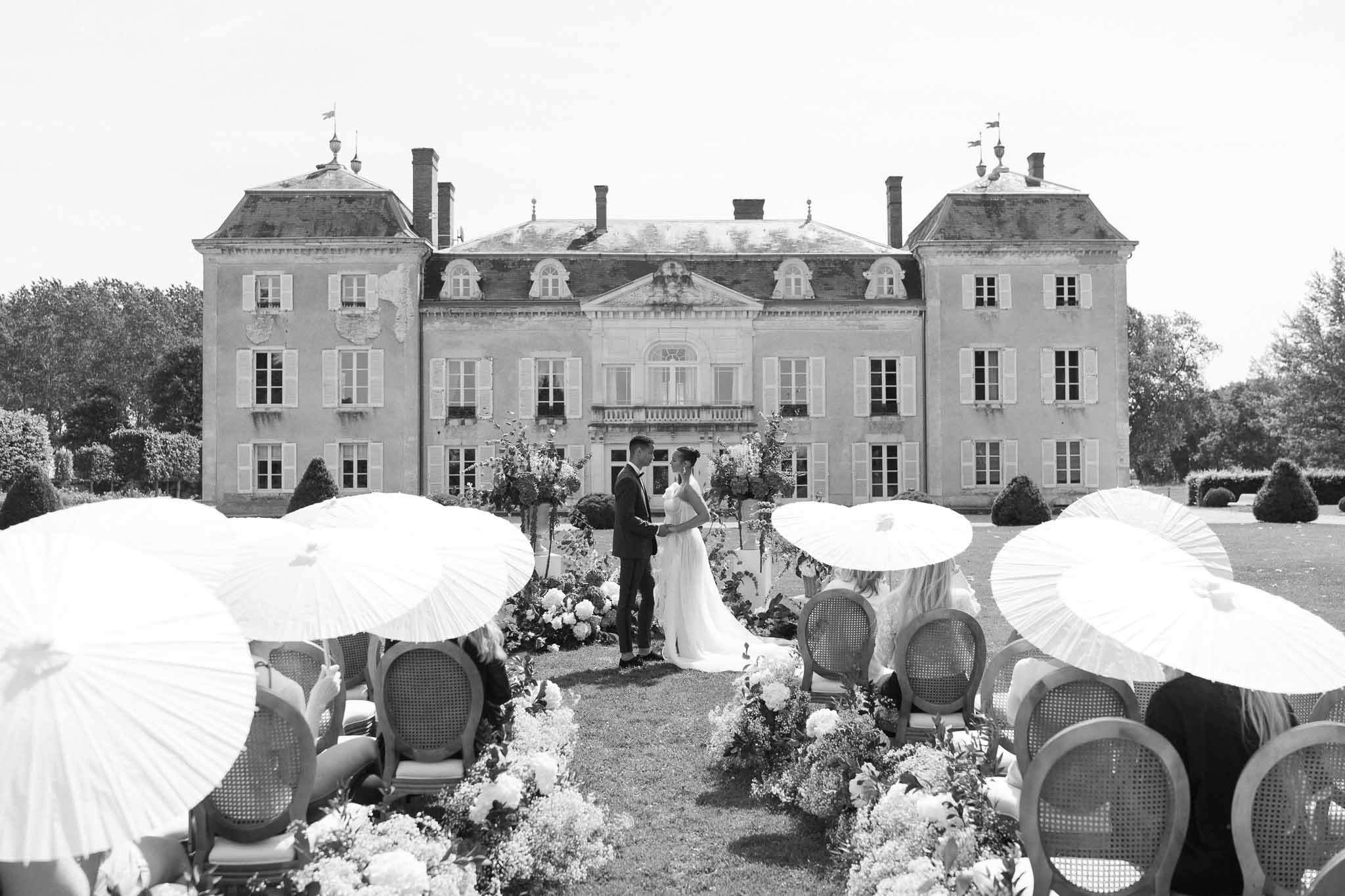 Outdoor wedding ceremony at château with guests holding parasols and formal gardens backdrop