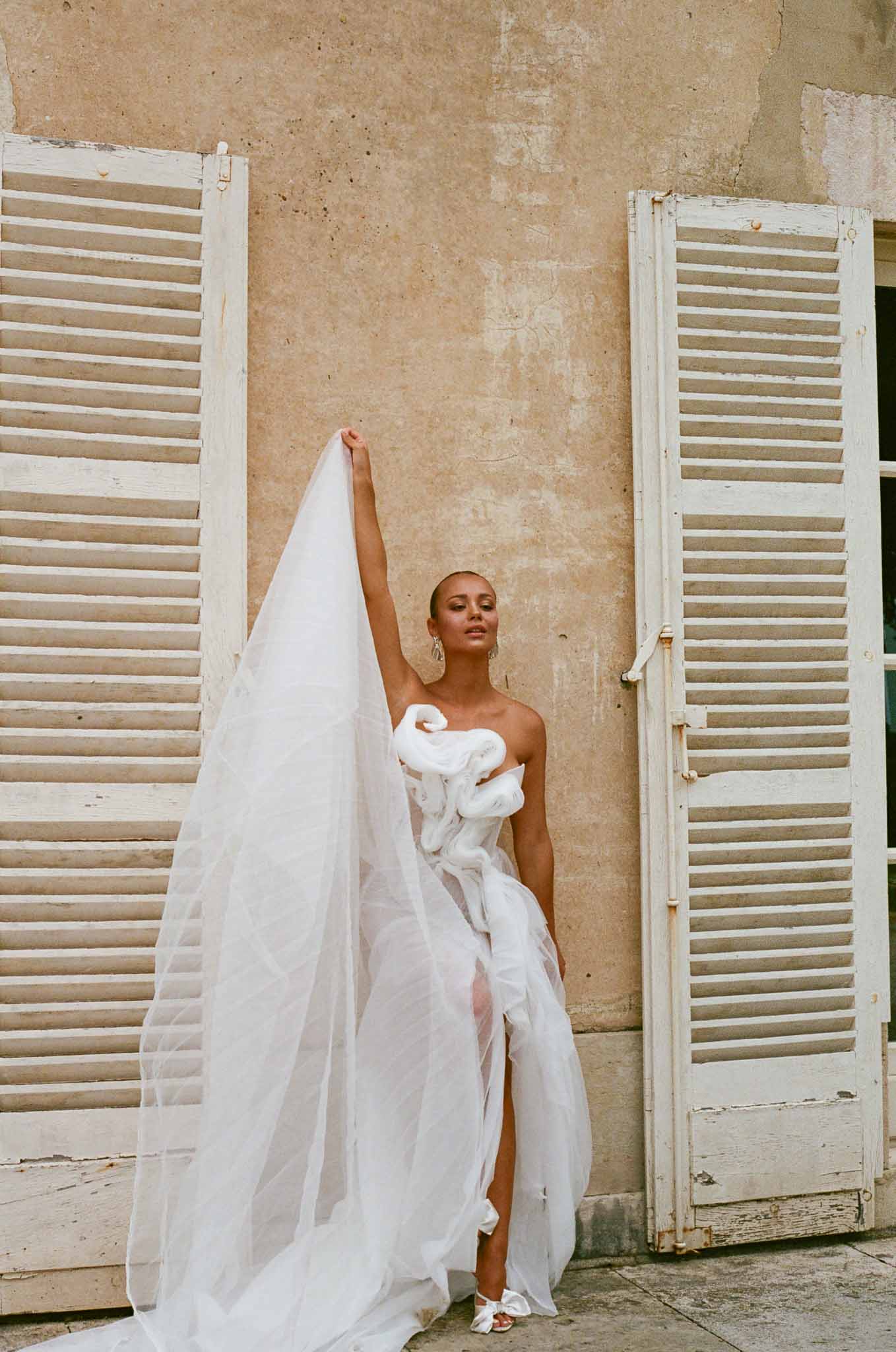 Bride in ivory wedding dress and veil posing against terracotta stone wall with vintage shutters