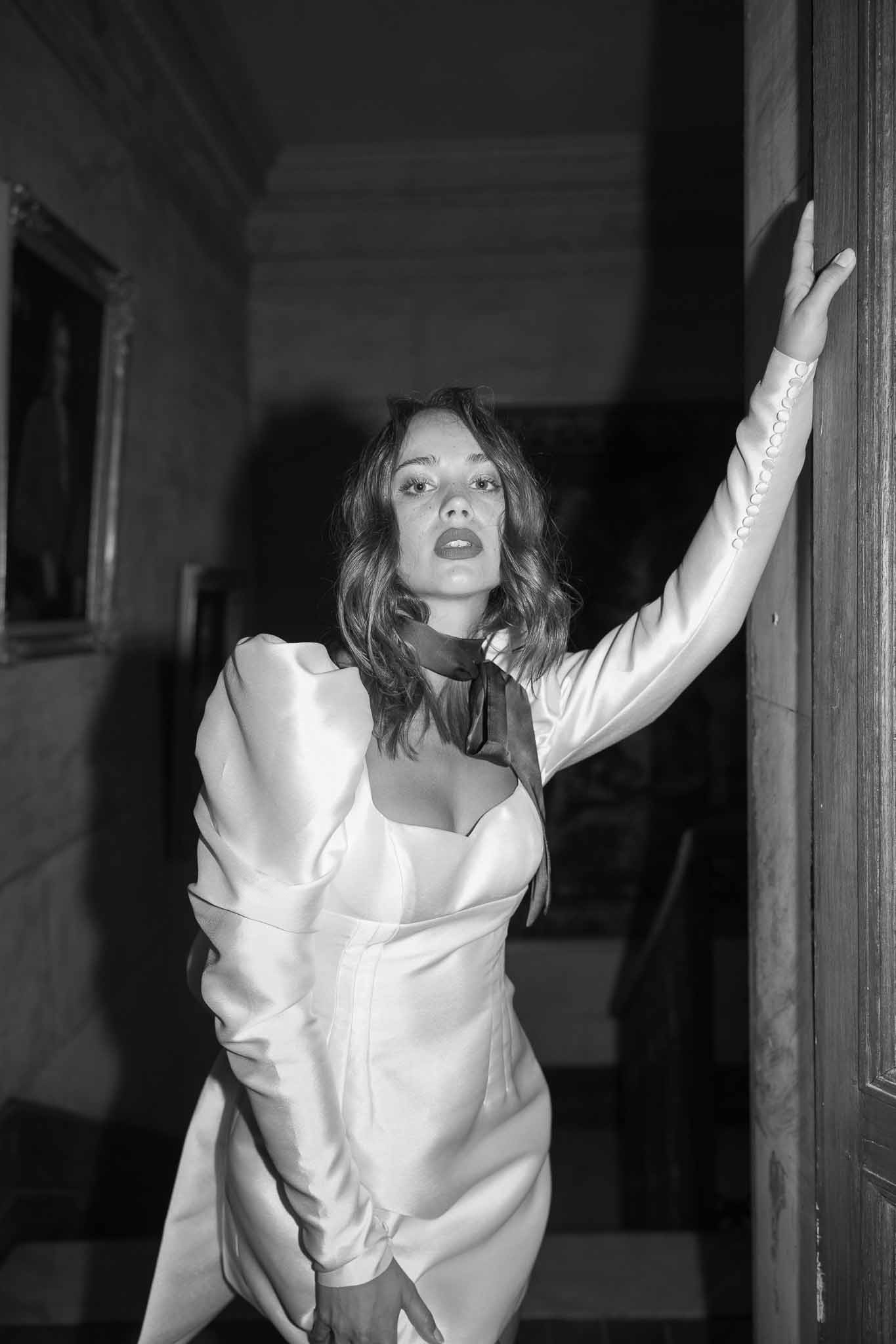 Bride in minimalist white dress with black ribbon posing against wooden doorframe in indoor hallway