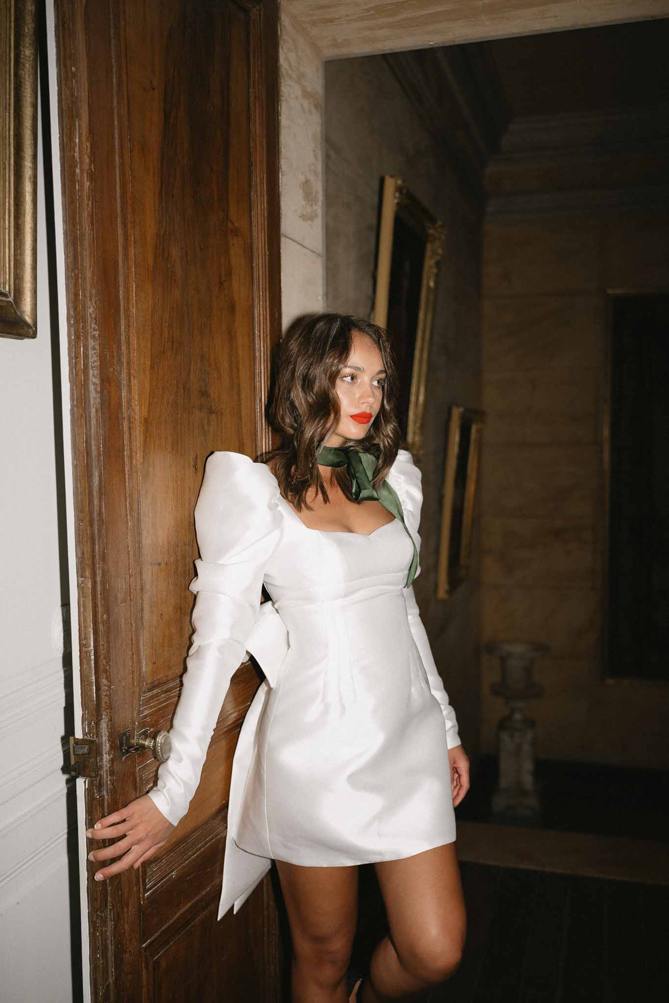 Bride in white mini dress with green ribbon posing in rustic stone corridor with wooden doors