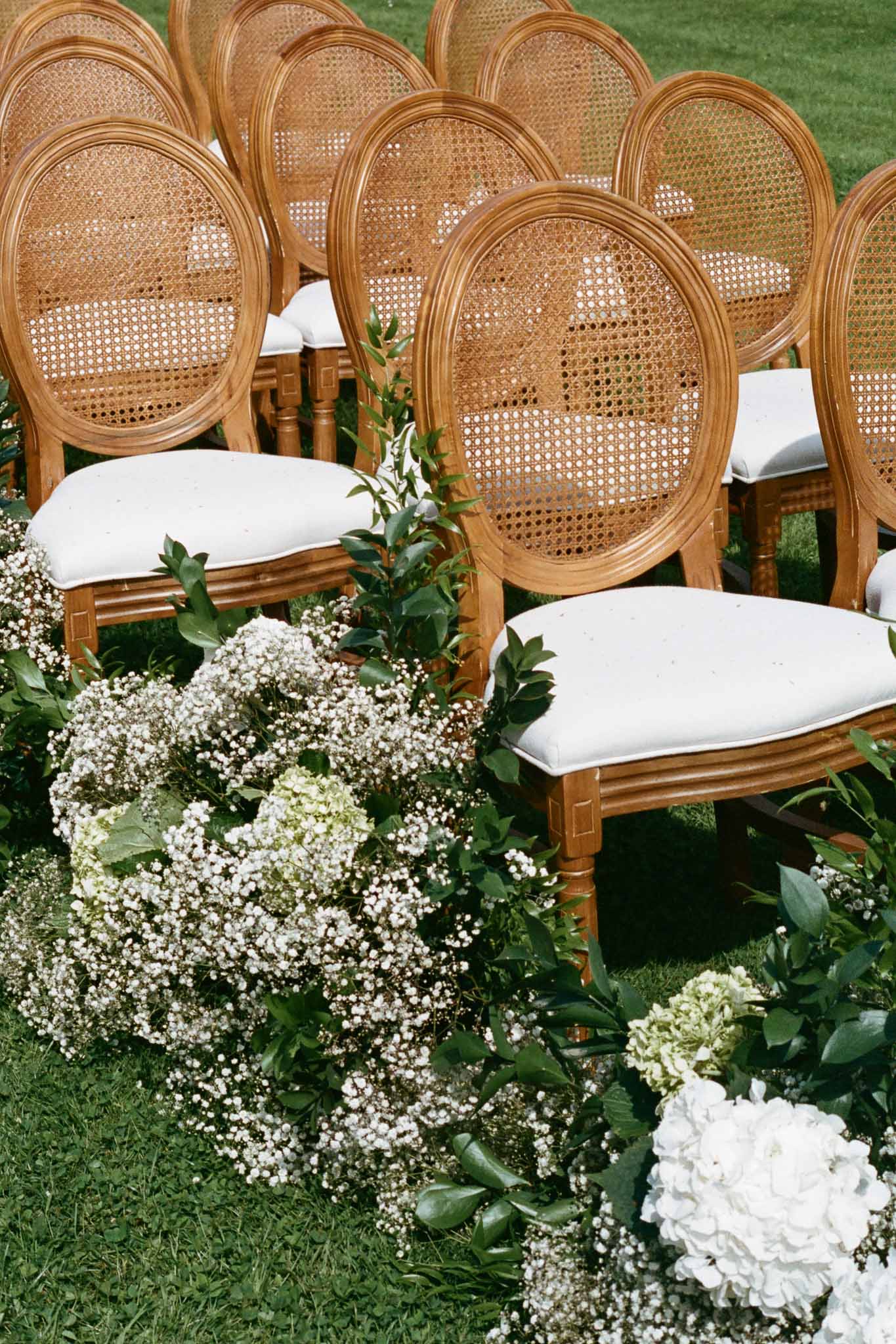 Wooden ceremony chairs with white florals arranged on outdoor lawn for wedding ceremony