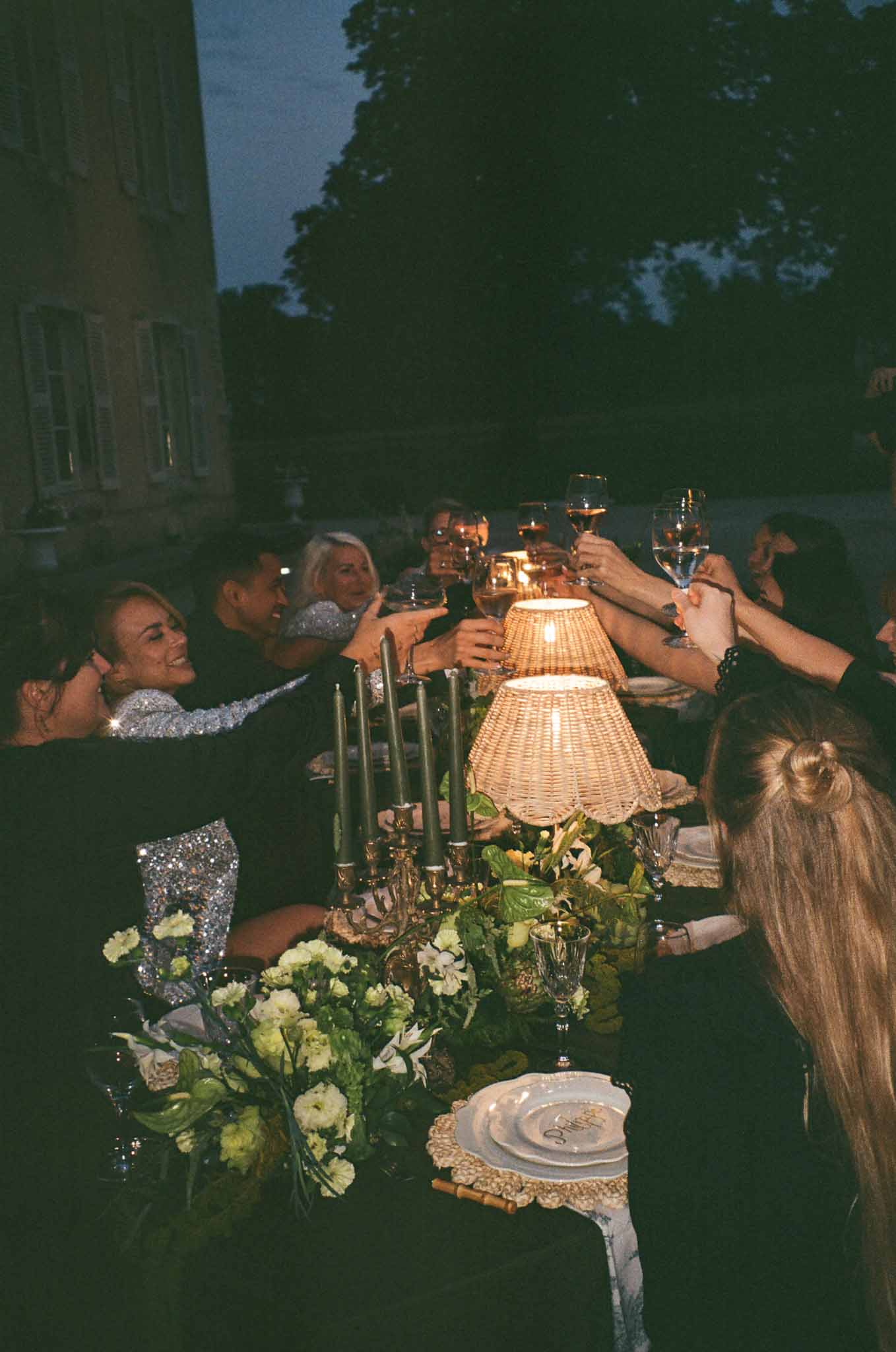 Evening wedding reception dinner with guests toasting at long table in garden courtyard setting