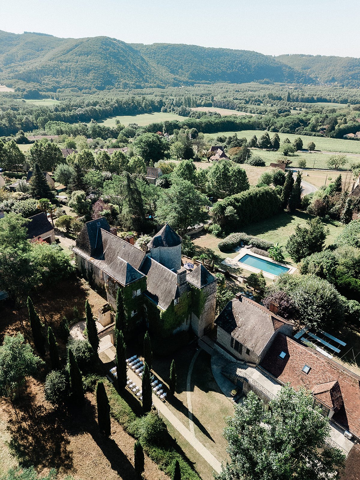 Aerial view of stone château wedding venue with pool surrounded by countryside and forested mountains