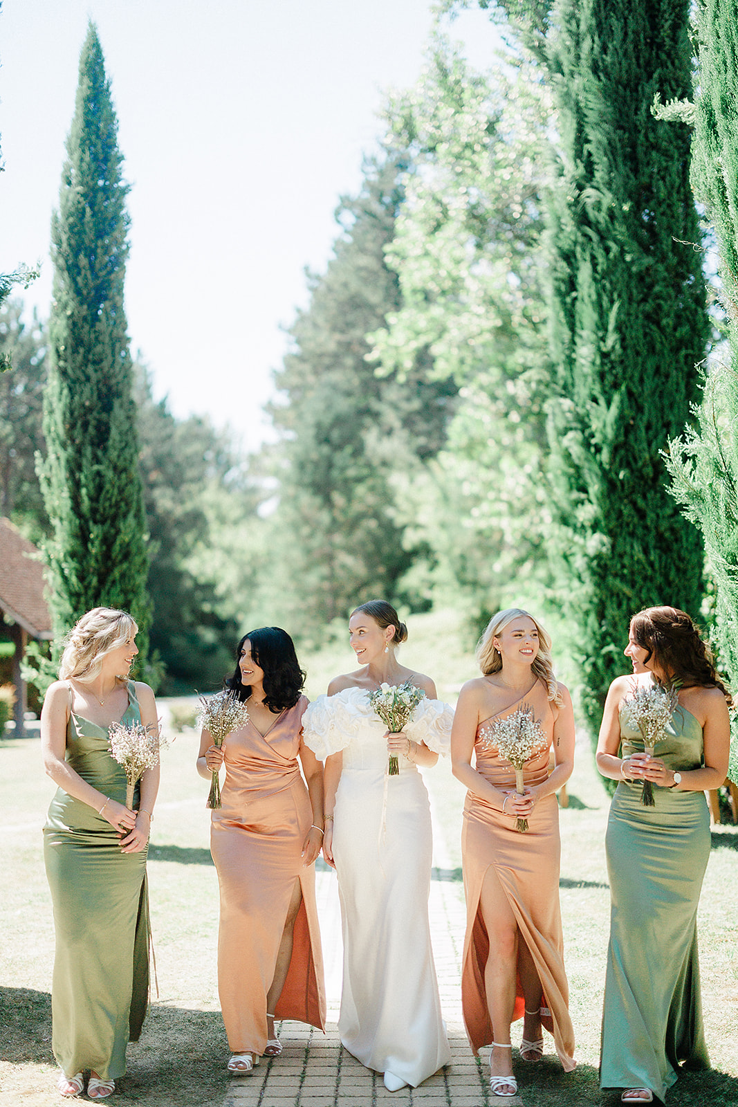 Bride and bridesmaids walking down cypress-lined pathway in formal garden setting
