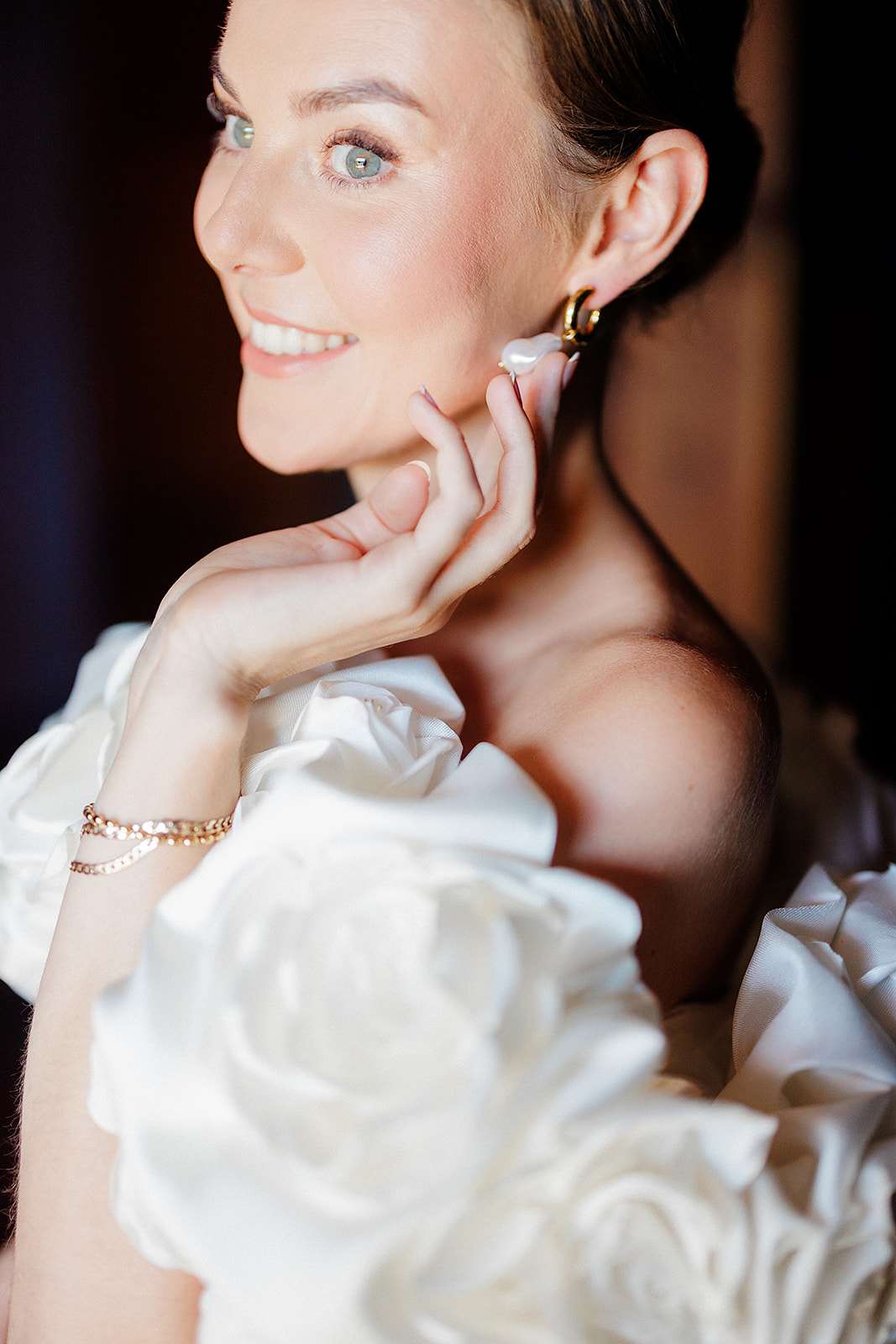 Bride in white off-shoulder dress with gold pearl jewelry smiling in profile portrait