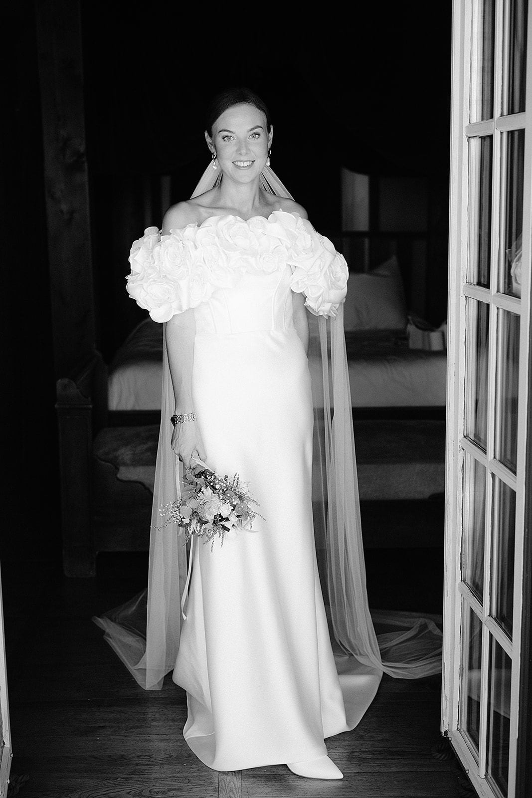 Bride in off-shoulder gown with floral detail standing in elegant hallway with French doors