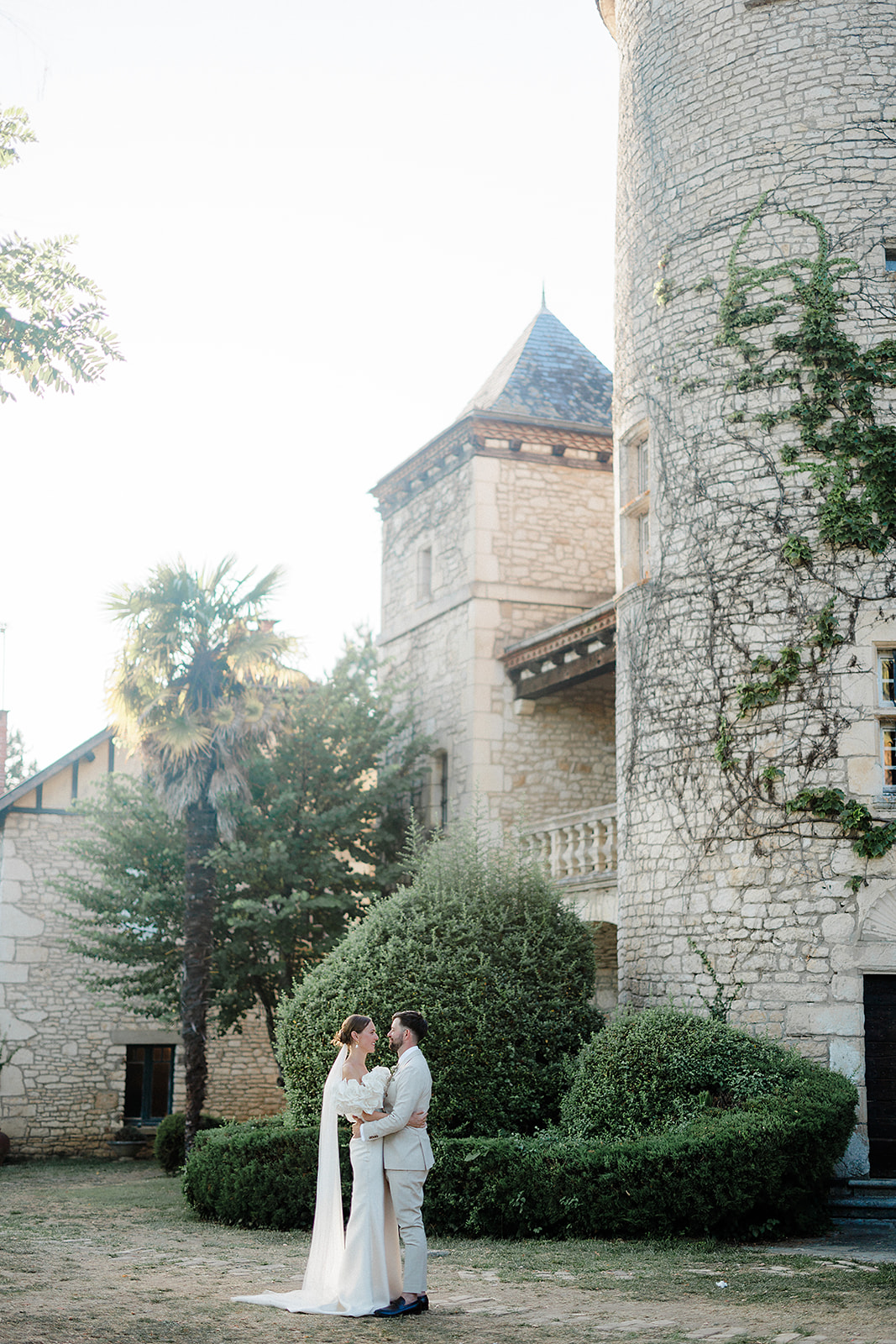 Bride and groom portrait in stone château courtyard with ivy-covered towers and manicured hedges