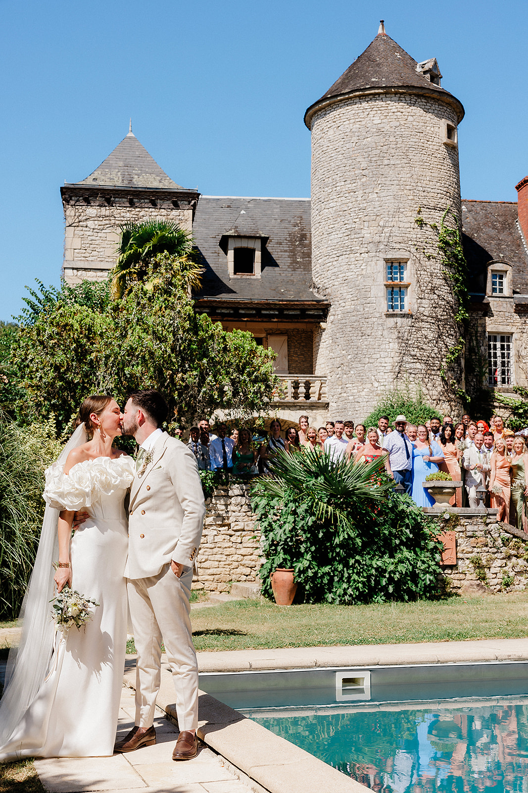 Bride and groom kissing poolside during reception at French château with guests on grounds