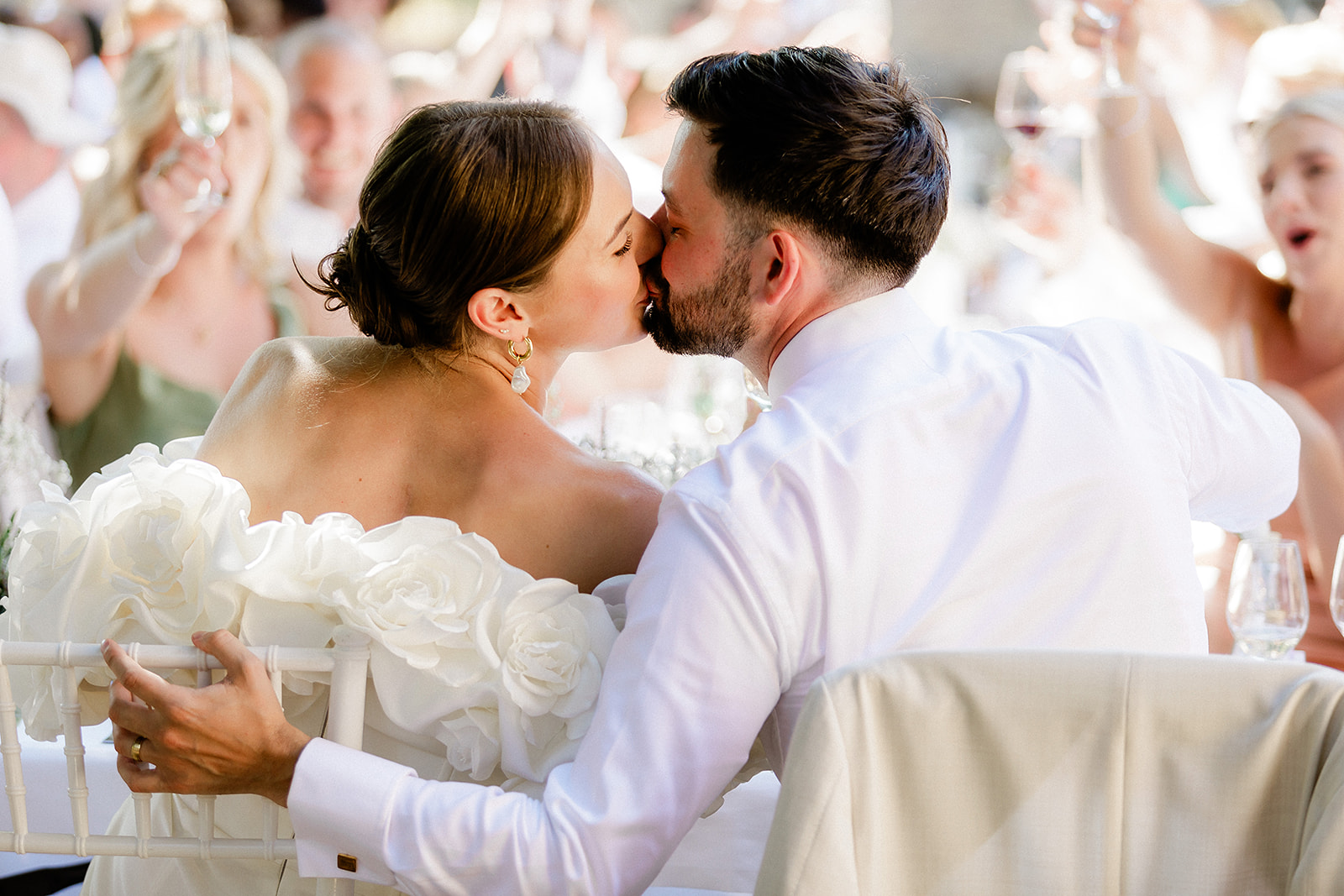 Bride and groom sharing first kiss at wedding reception with guests cheering in background