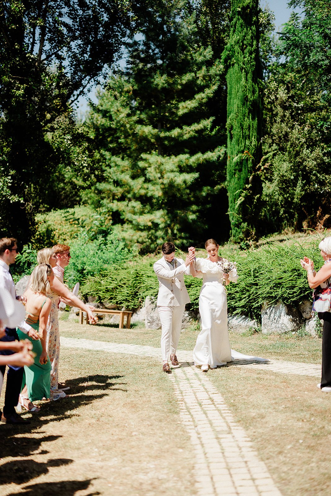 Bride and groom walking down ceremony aisle with guests in garden setting with cypress trees