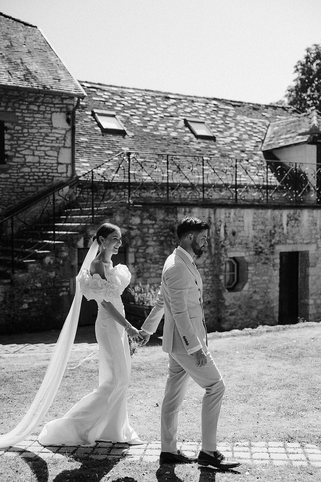 Bride and groom walking hand-in-hand in historic stone courtyard with slate roofs and iron footbridge