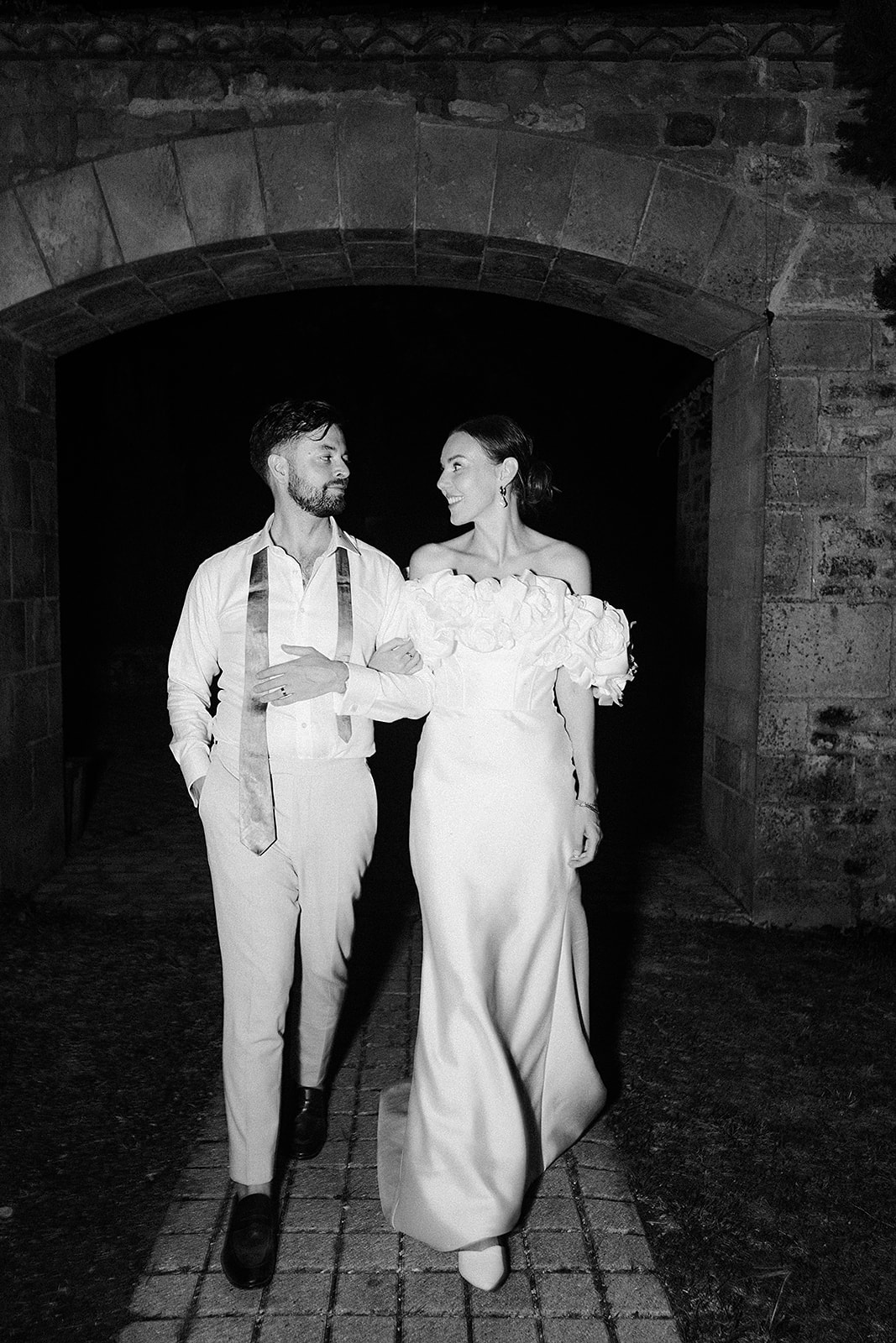 Bride and groom walking through historic stone archway in black and white portrait