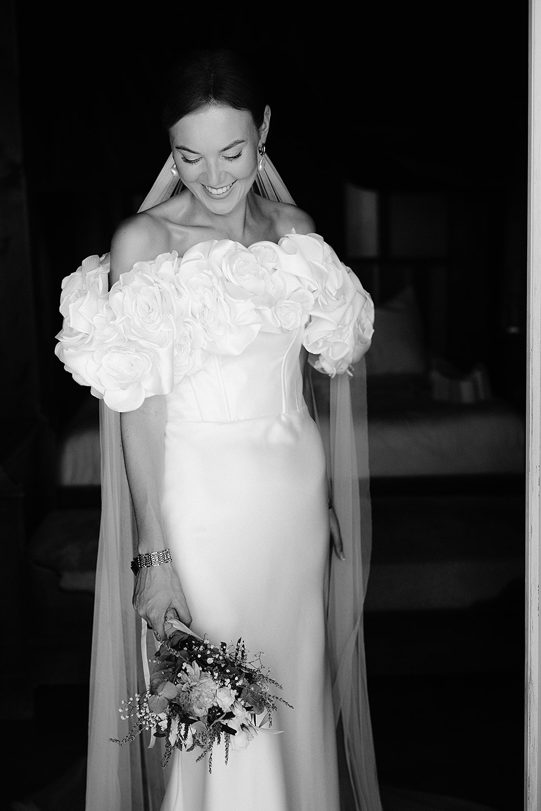 Black and white bridal portrait with rose shoulder arrangement and flowing veil against dark background