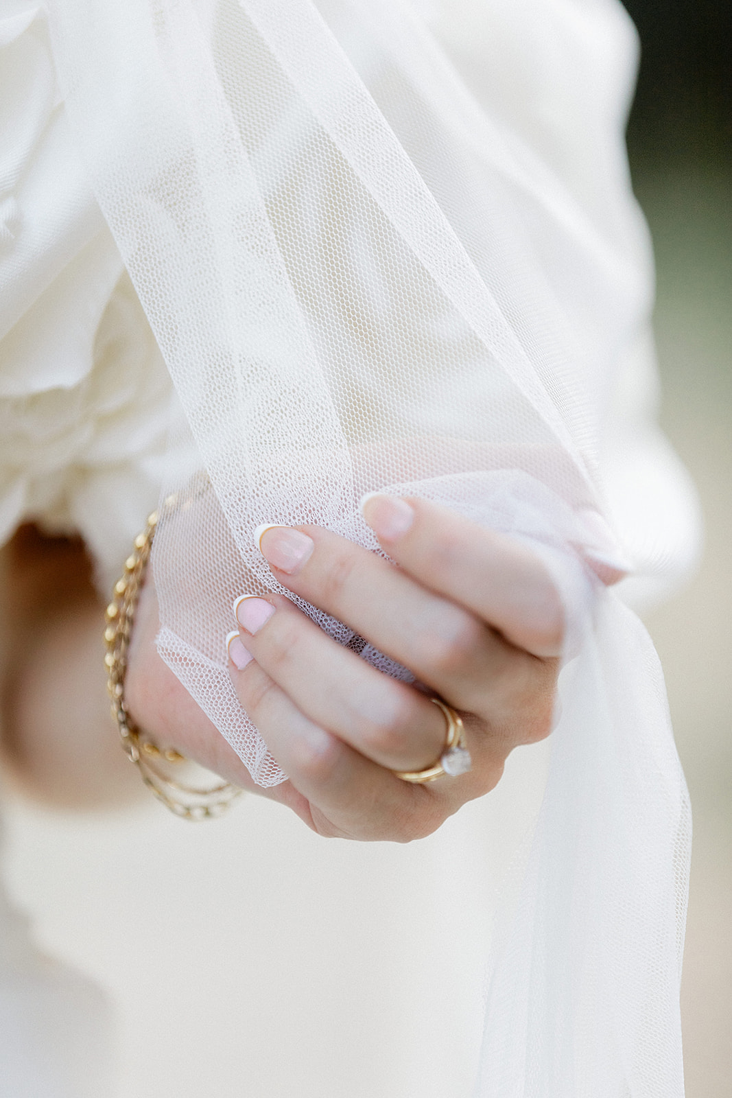 Close-up of bride's hand holding ivory tulle wedding veil with gold jewelry and manicured nails