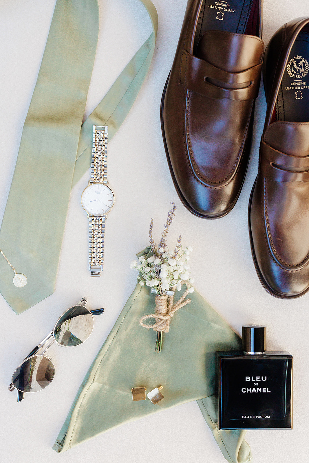 Groom's wedding accessories flat lay with sage tie, brown shoes, watch, and boutonnière on white background