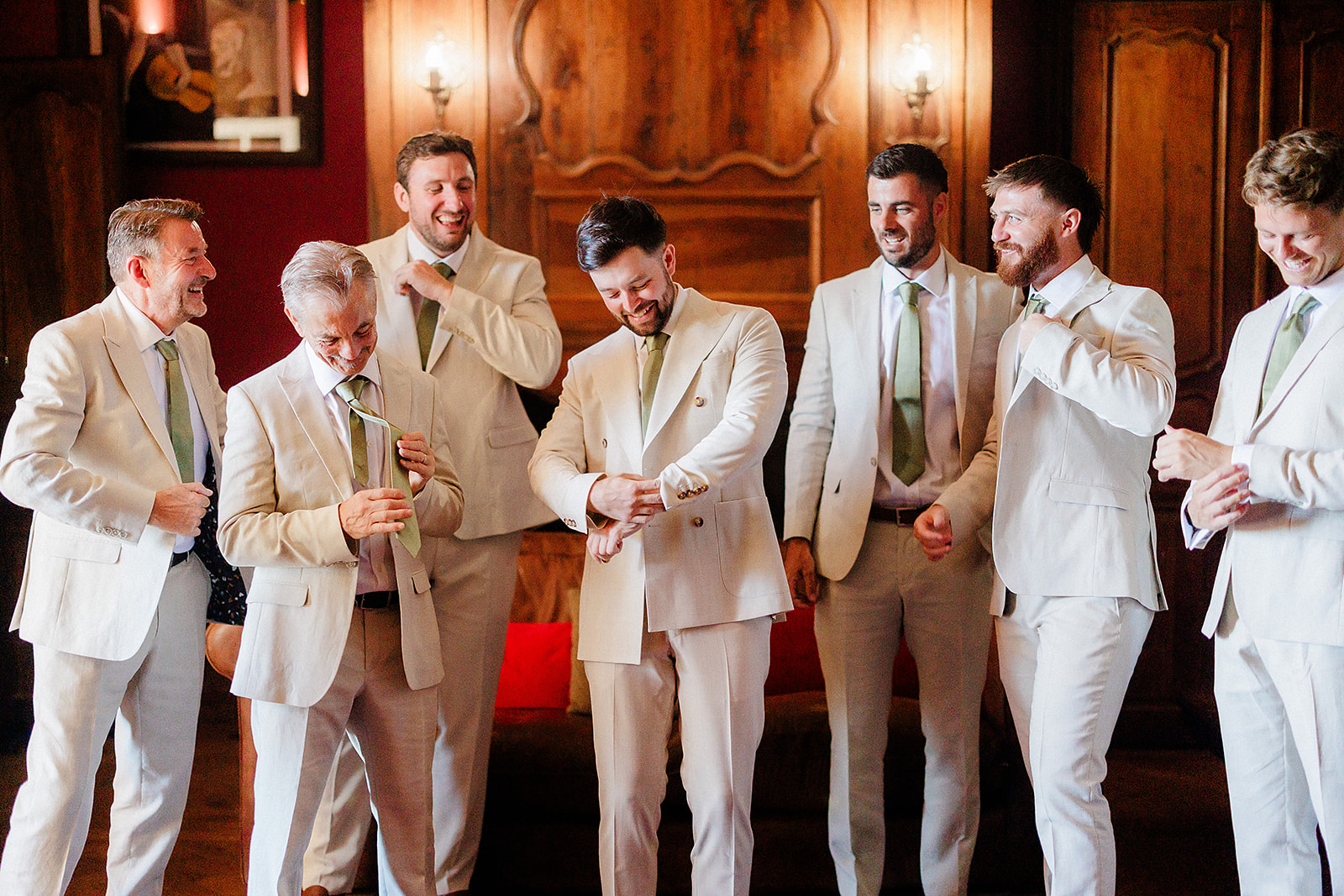 Groom and groomsmen laughing together during wedding reception in ornate interior with burgundy walls