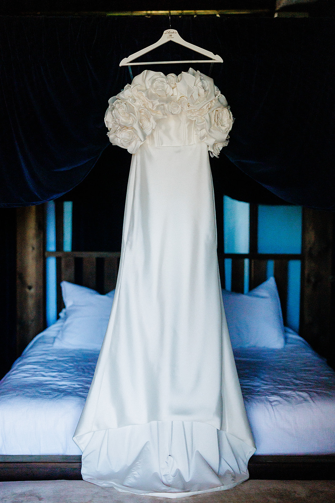 Ivory wedding dress with three-dimensional roses hanging in elegant bedroom with navy curtains
