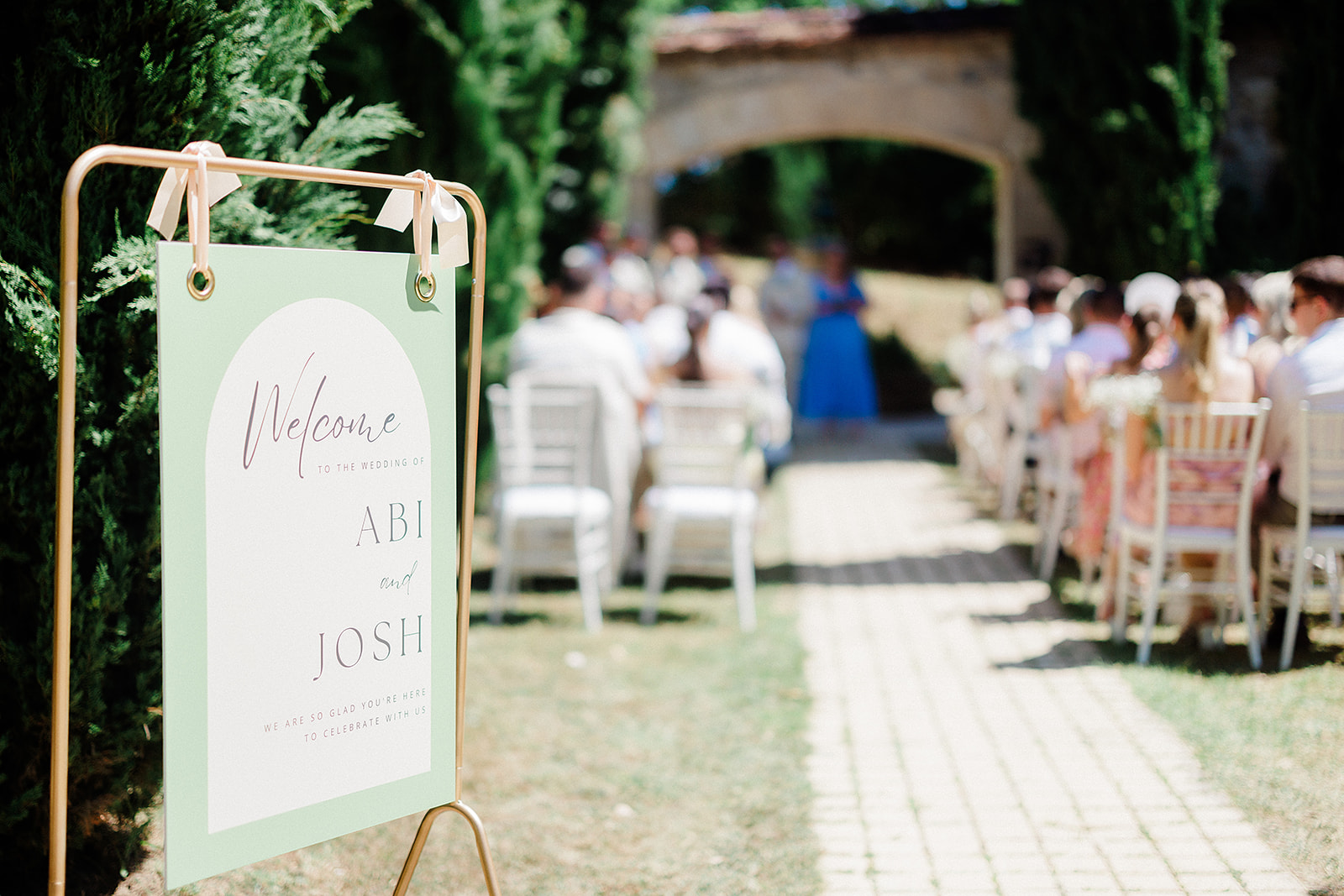Wedding ceremony welcome sign with guests seated in outdoor courtyard with stone archway