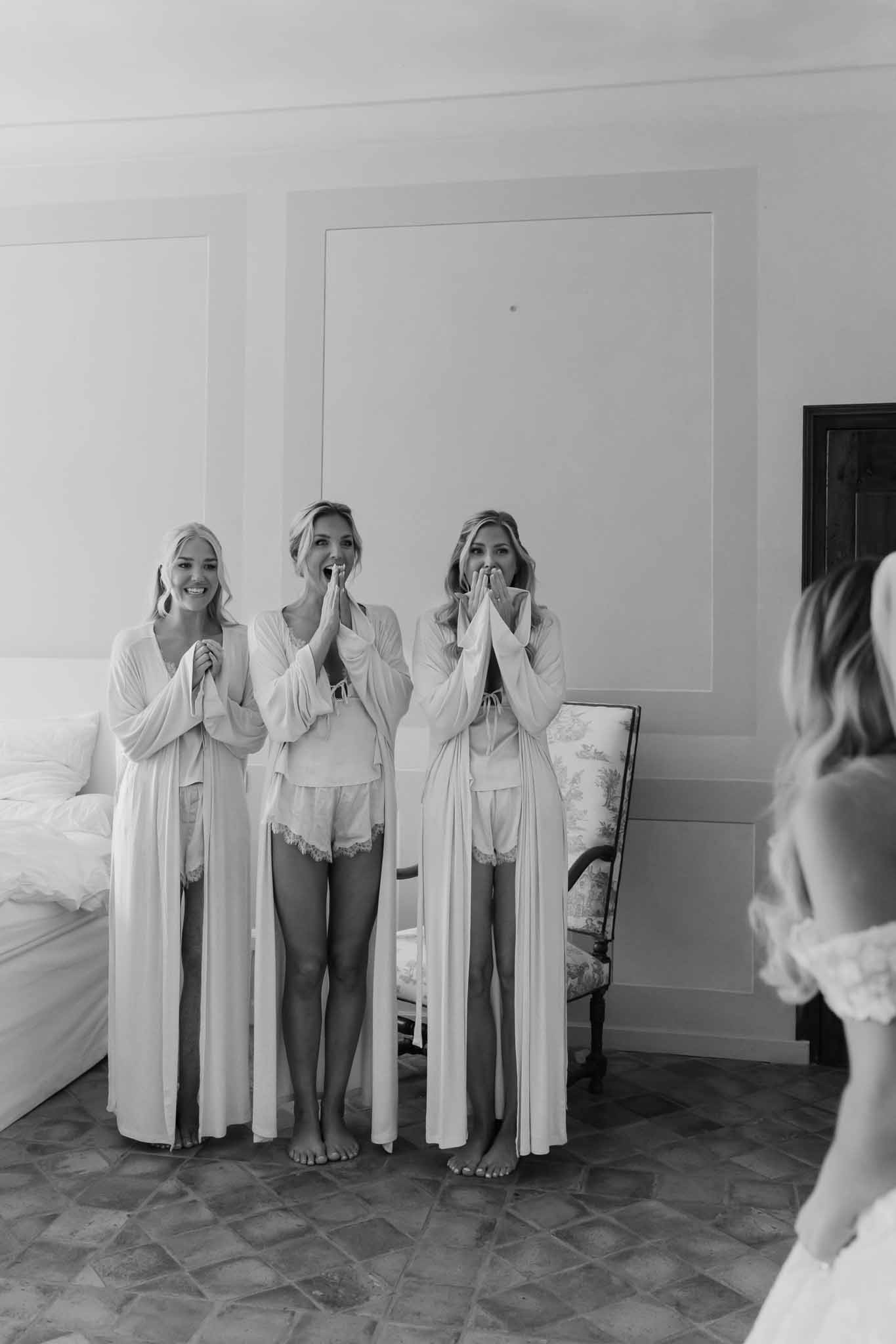 Bridesmaids in cream robes reacting with joy during getting ready moments in bridal suite