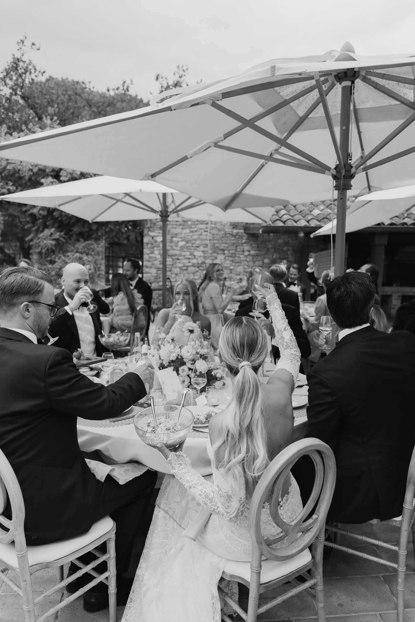 Reception dinner with bride at banquet table during toasts at outdoor stone venue with umbrellas