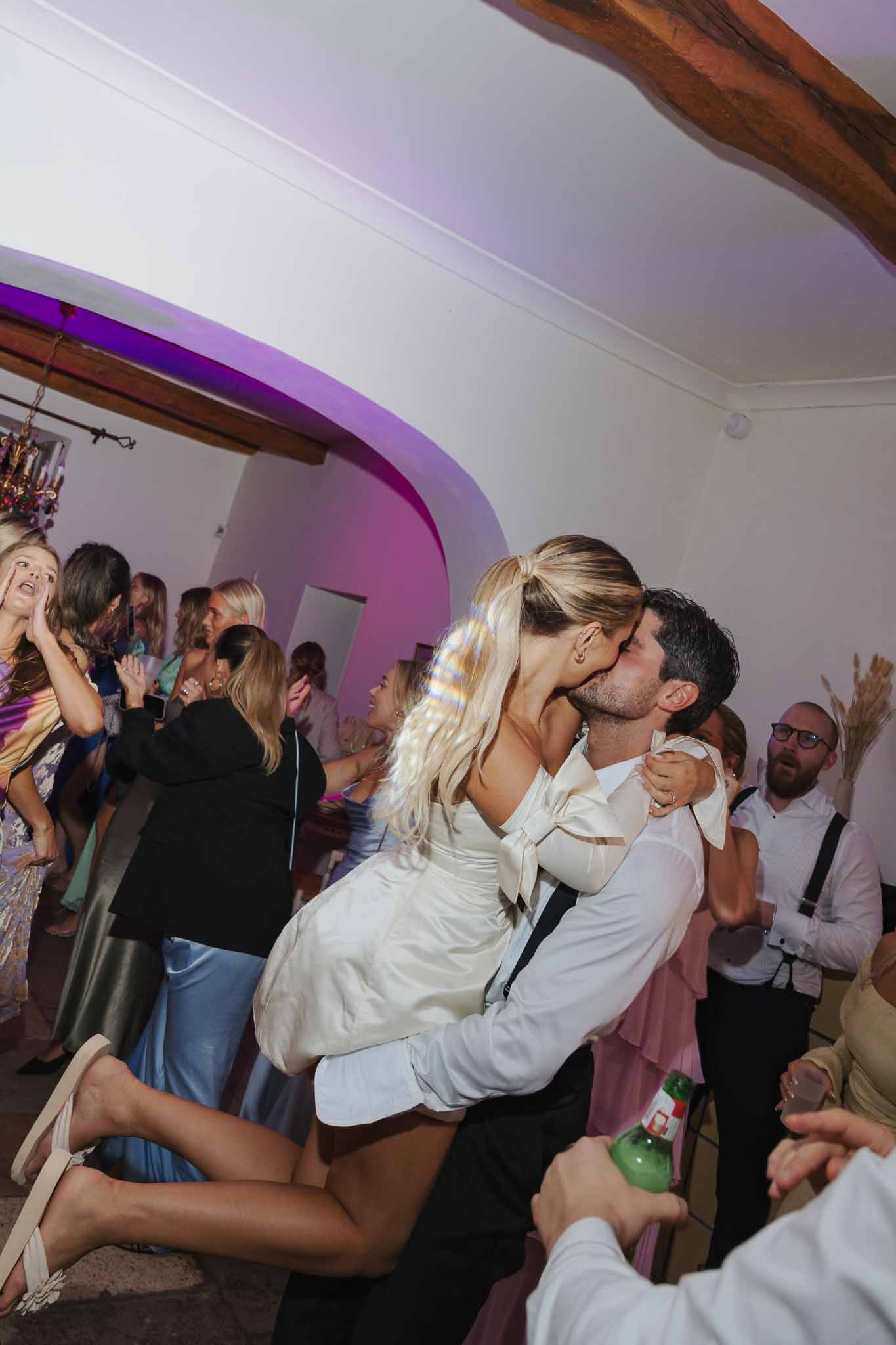 Groom lifting and kissing bride during reception dance with cheering guests in rustic venue with exposed beams
