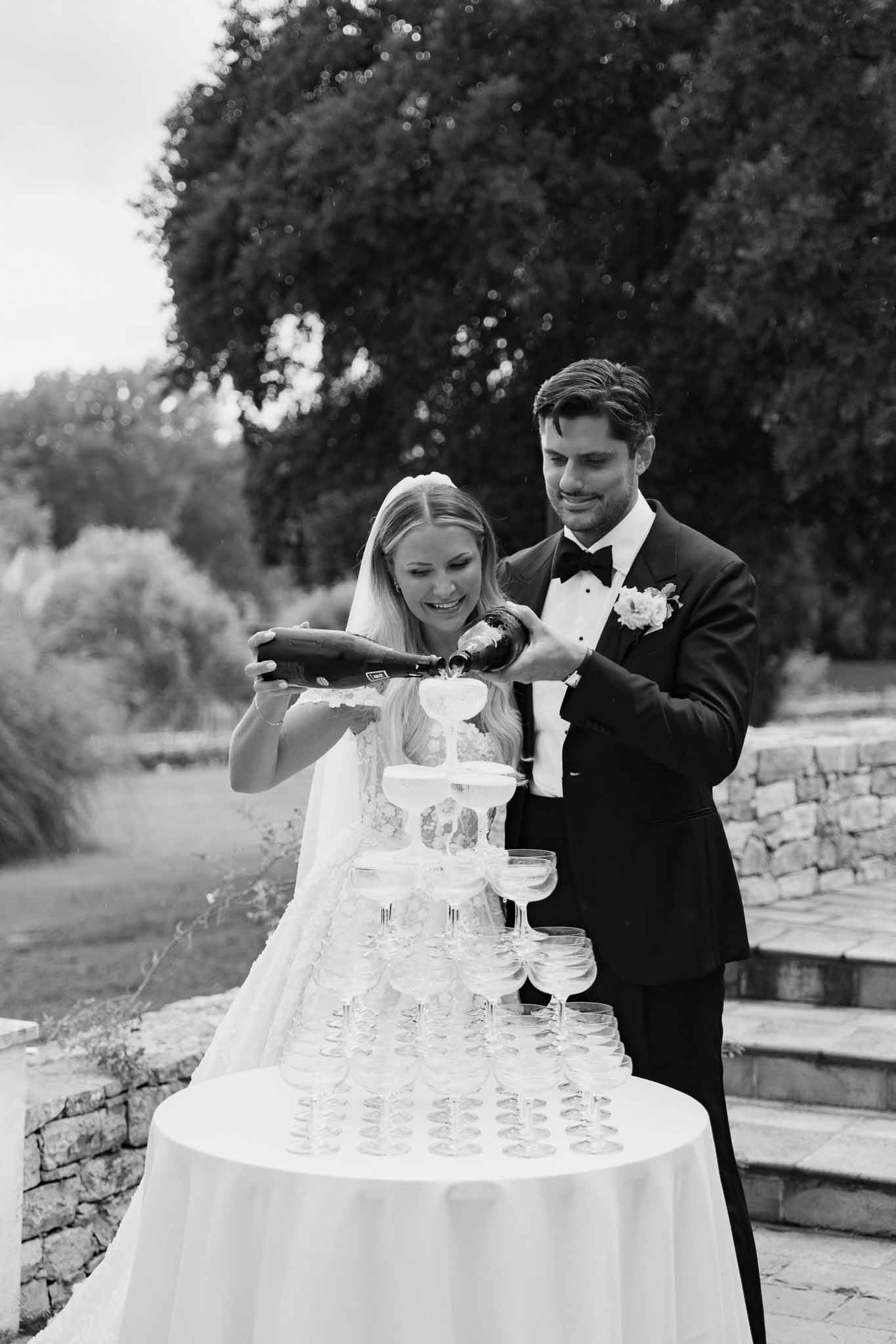 Bride and groom pouring champagne tower together during outdoor wedding reception in garden setting