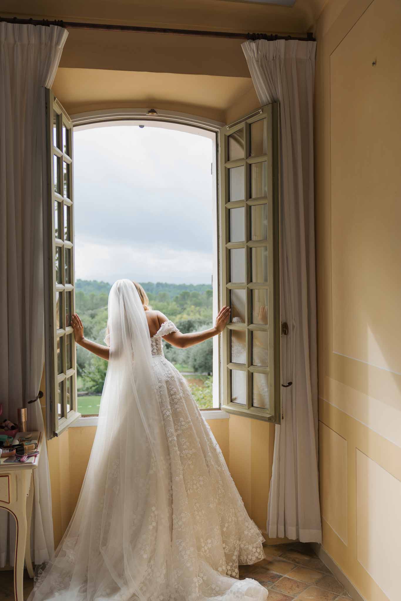 Bride in ivory lace dress and veil standing at French doors overlooking countryside landscape