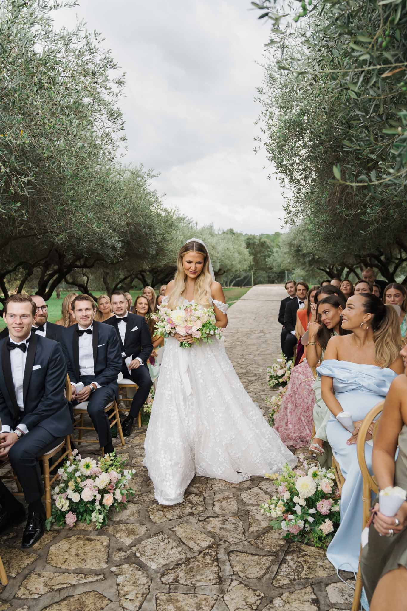Bride walking down outdoor aisle during wedding ceremony in olive grove with seated guests