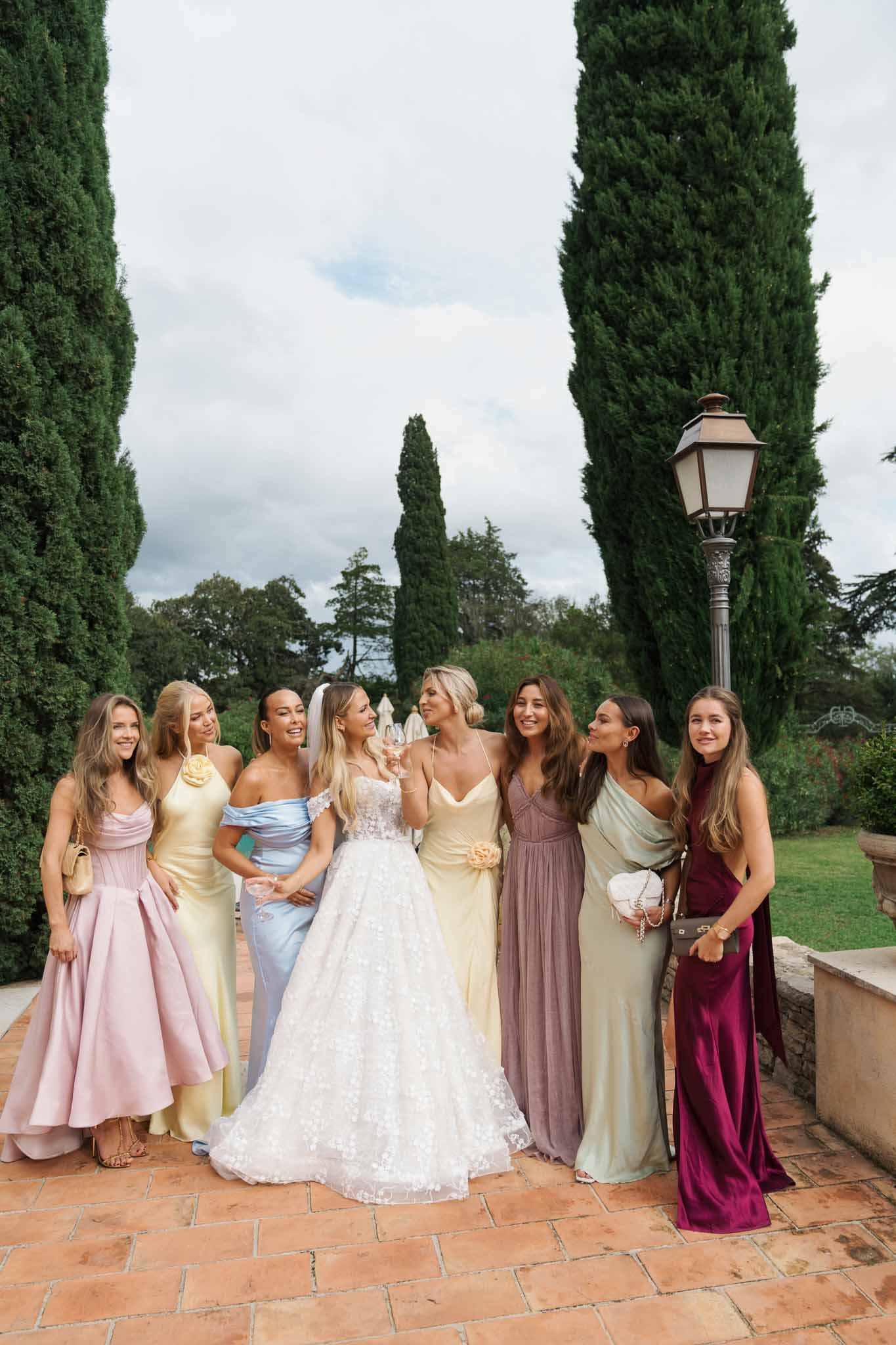 Bride with eight bridesmaids in pastel gowns on cypress-lined pathway at garden estate