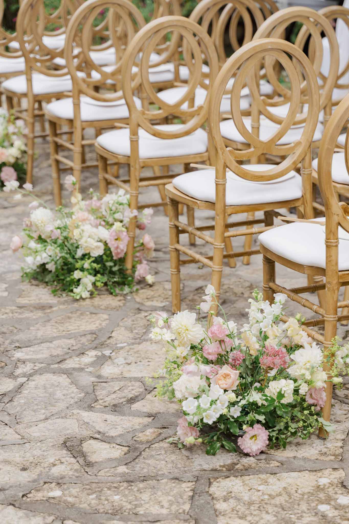 Wedding ceremony seating arrangement with wooden chairs and blush floral arrangements on stone courtyard