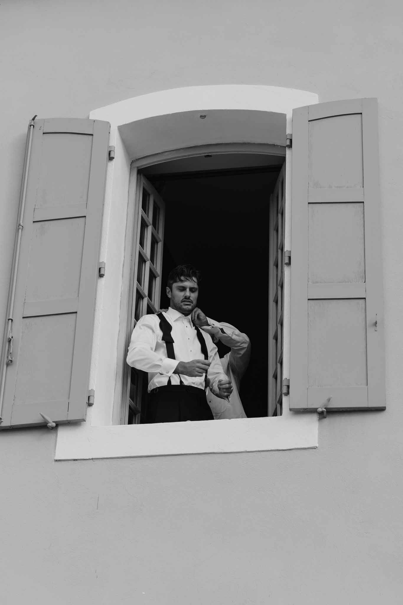 Groom adjusting clothing in window with white shutters during wedding preparation
