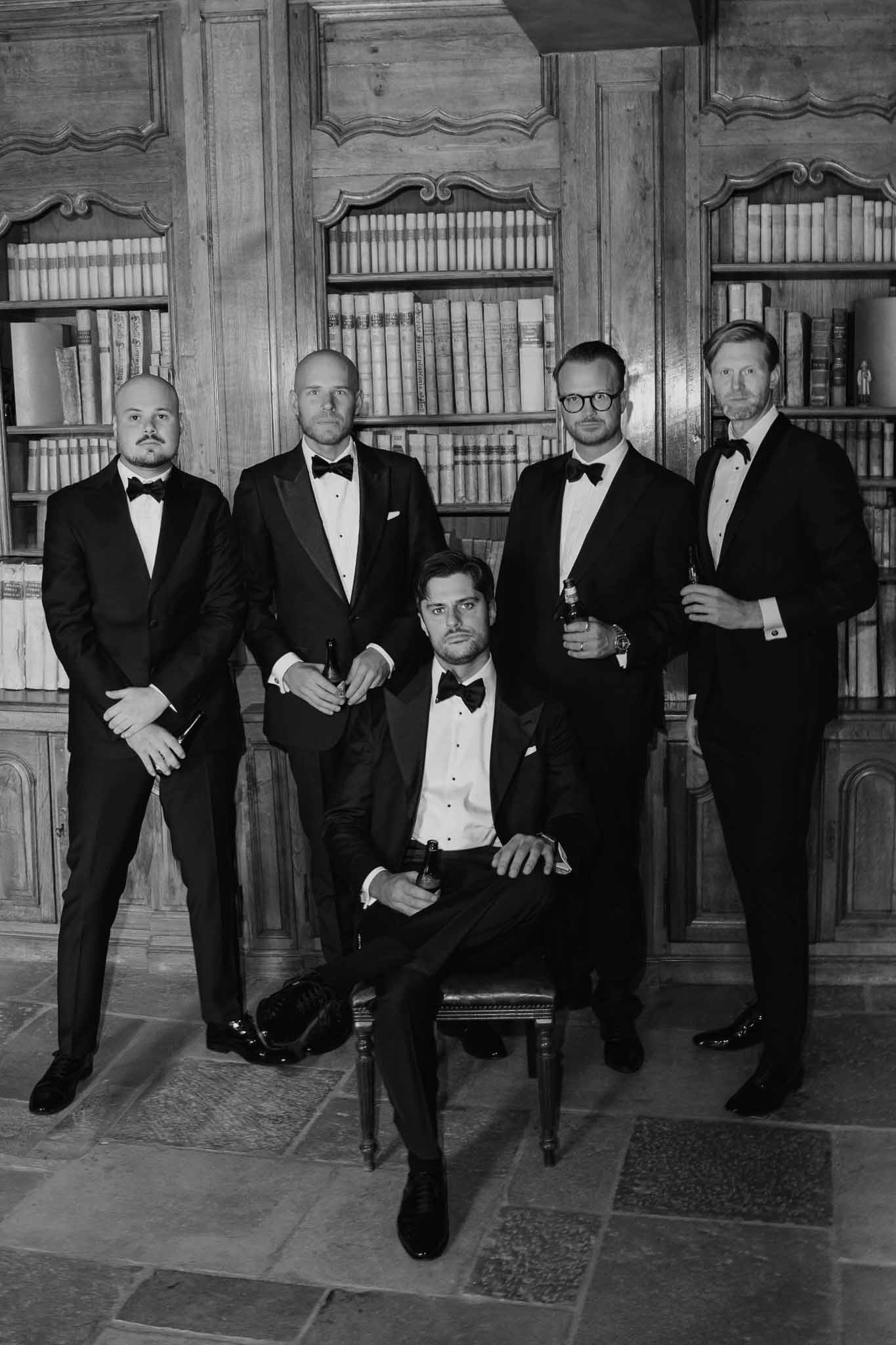 Five groomsmen in black tuxedos posed in elegant library with wooden bookshelves