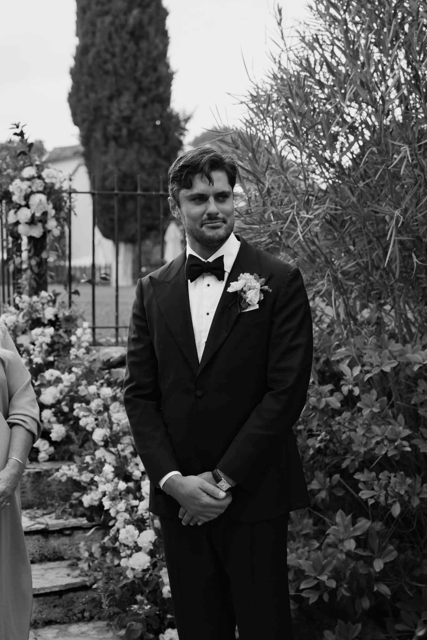 Groom in black tuxedo waiting at ceremony altar in Mediterranean garden setting