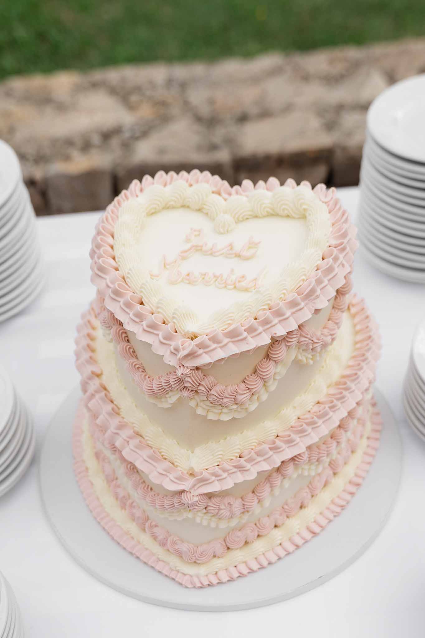 Three-tier heart-shaped wedding cake with pink and ivory frosting on white stand at outdoor reception