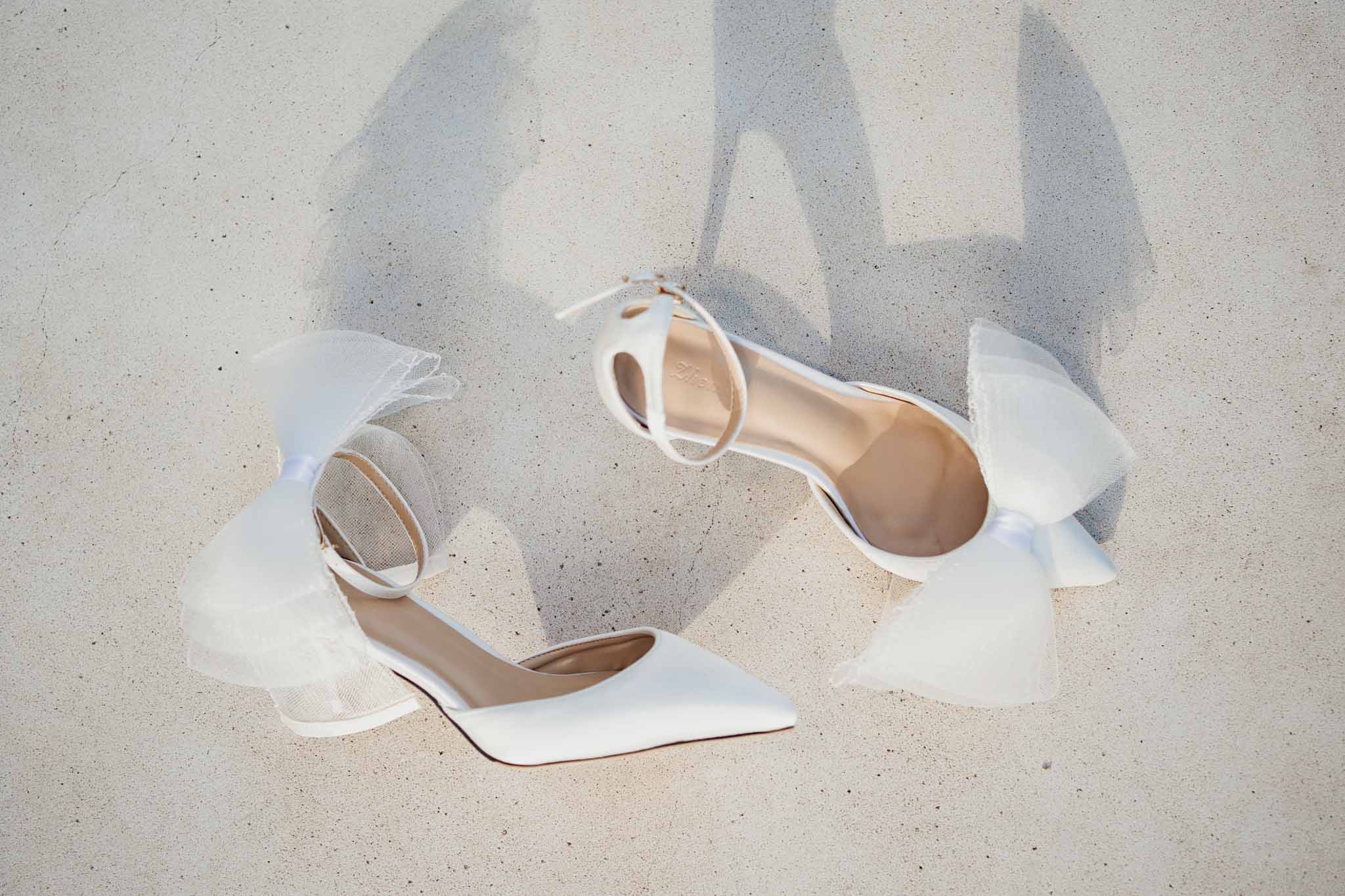 White bridal shoes with ankle straps and bow details on sand at beach wedding venue
