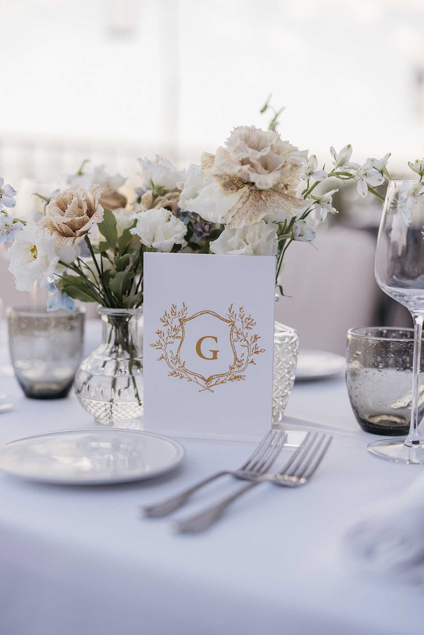Reception table setting with gold monogram place card and white floral centerpieces at wedding venue
