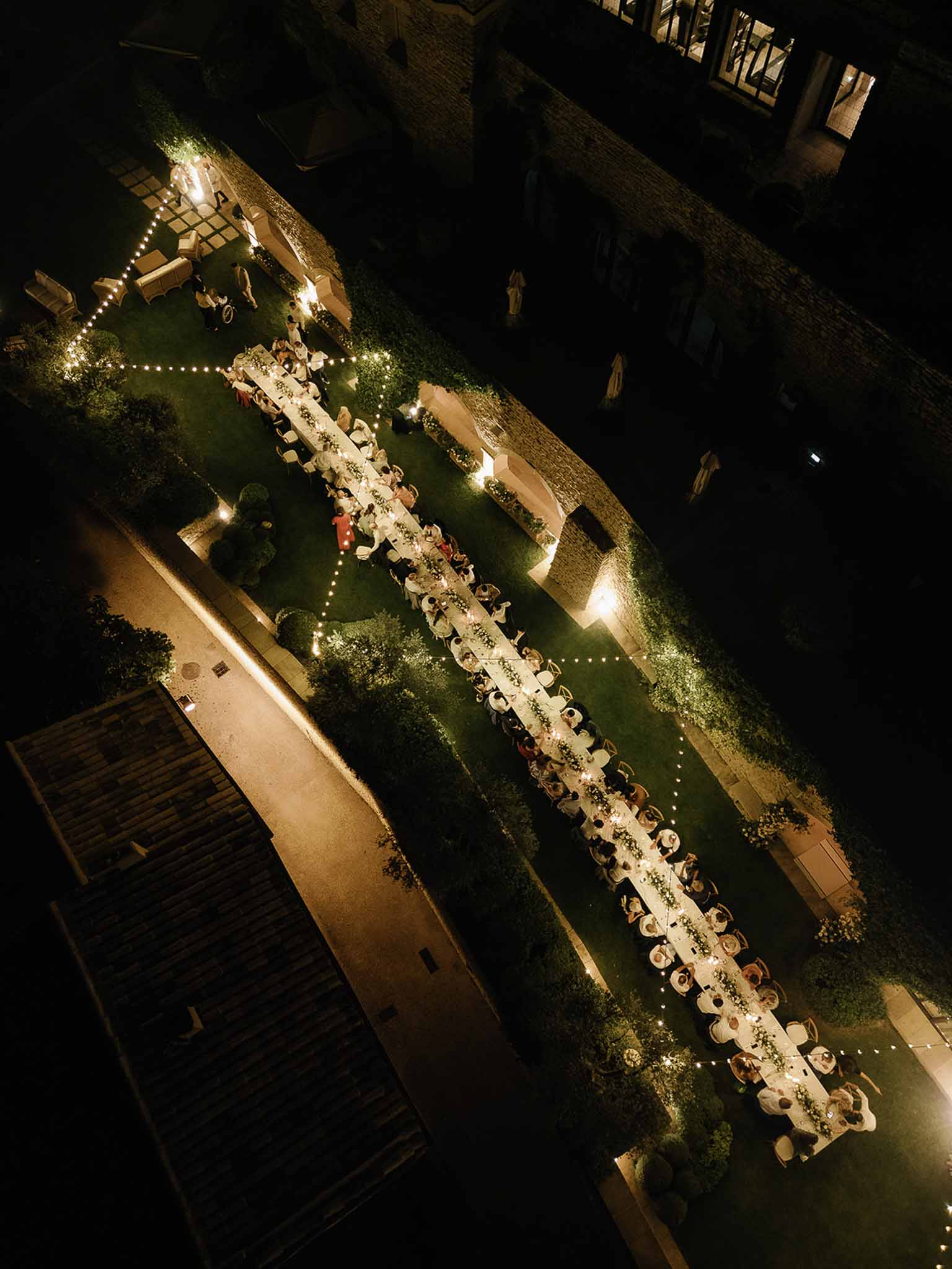 Aerial view of outdoor reception dinner in historic stone courtyard with string lights and long dining table