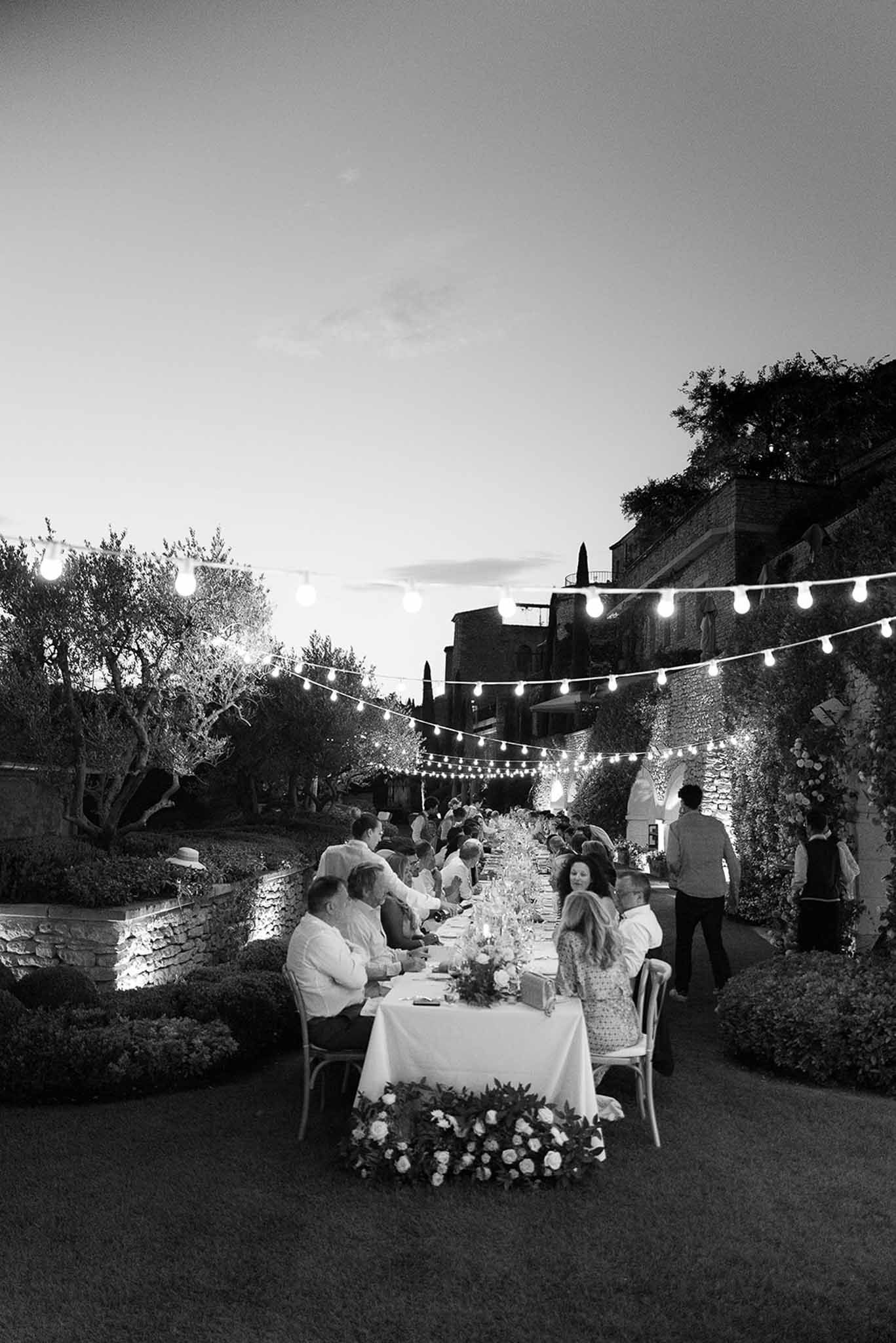 Reception dinner with guests at long table under bistro lights at outdoor Tuscan stone venue