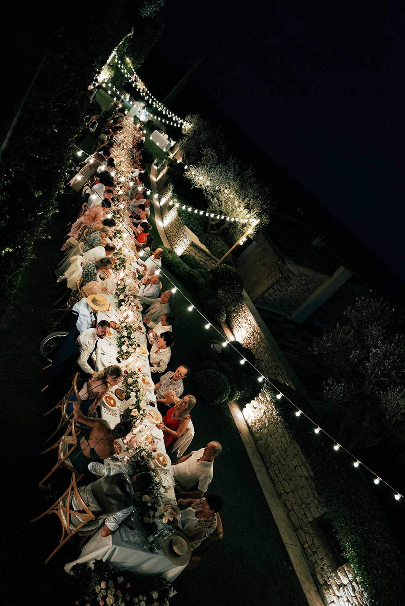 Aerial view of outdoor reception dinner with long banquet table in stone courtyard at night