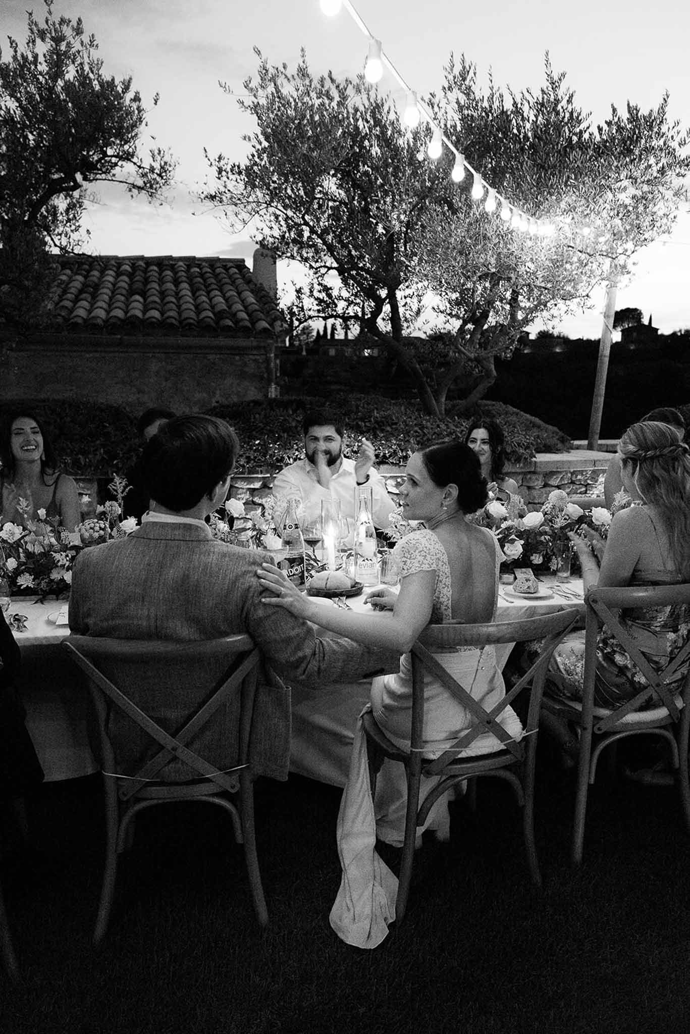 Wedding guests dining and laughing at outdoor reception table with Mediterranean stone venue backdrop