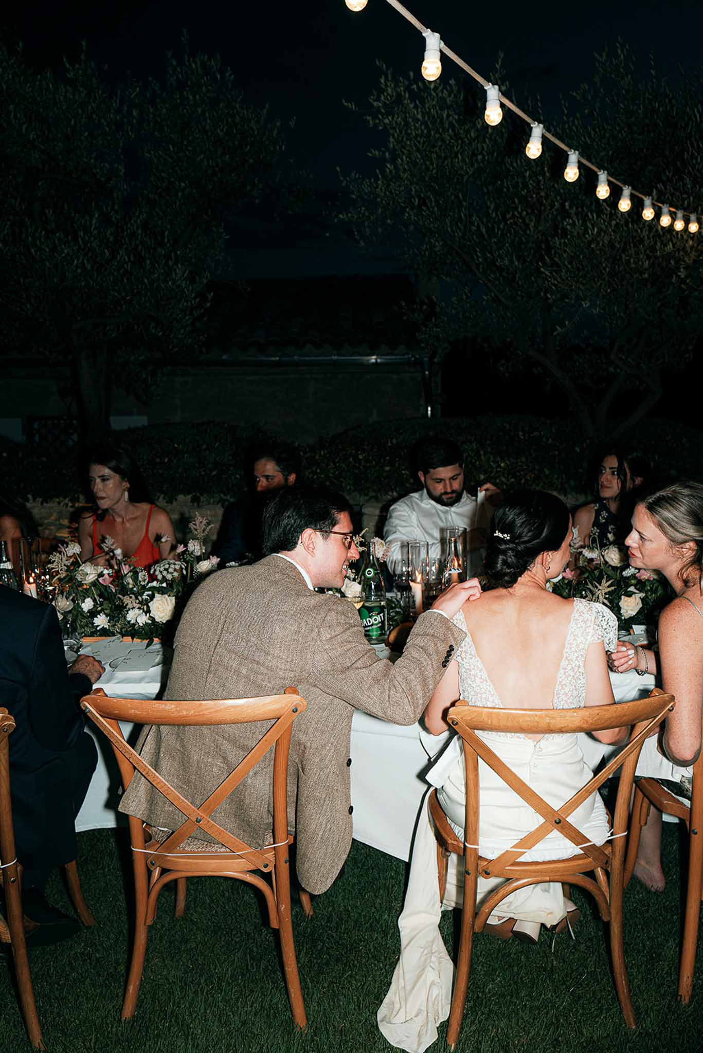 Outdoor evening reception dinner with guests at long table under string lights in garden setting