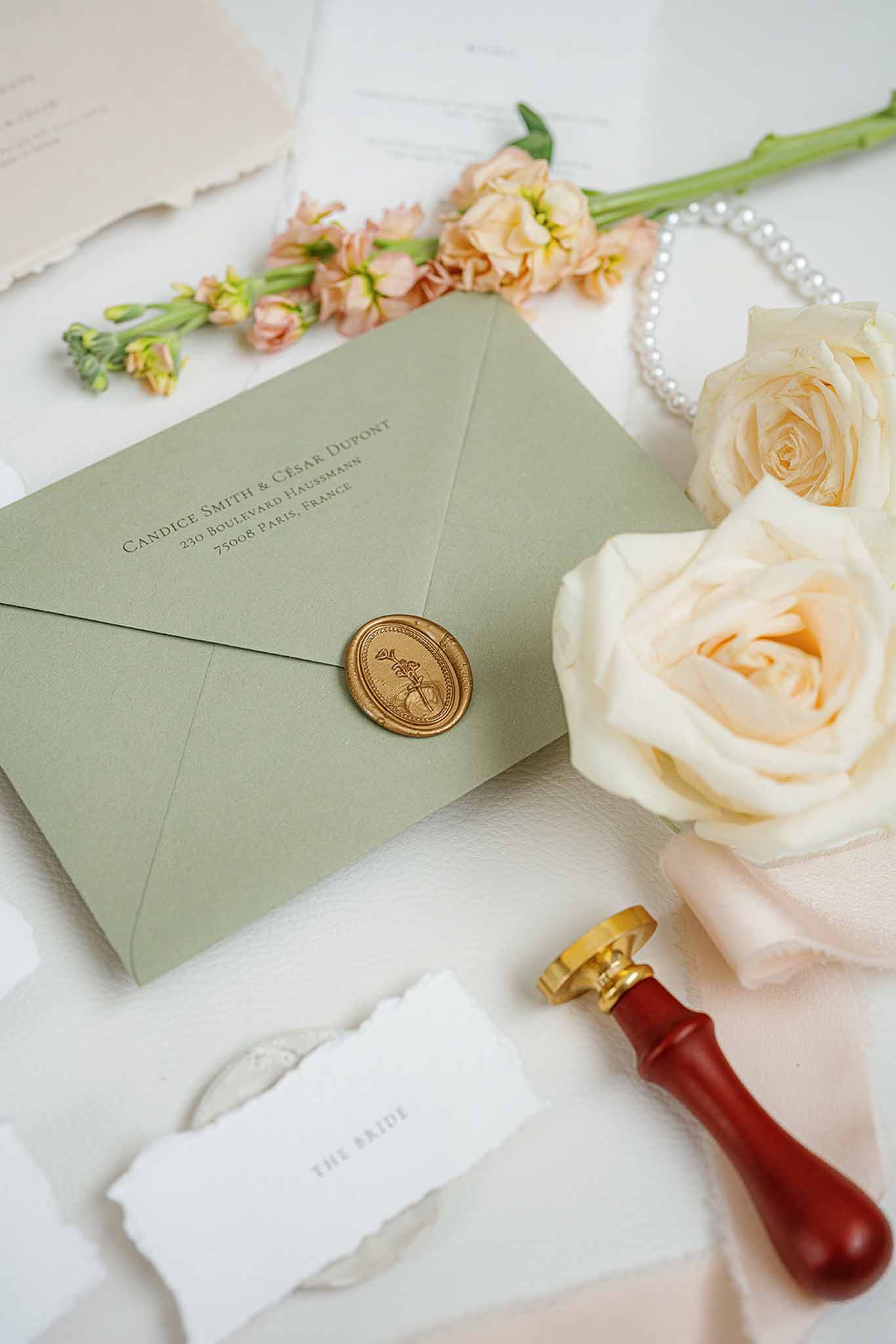 Wedding stationery flat lay with sage envelope, peach roses, and gold wax seal on white surface