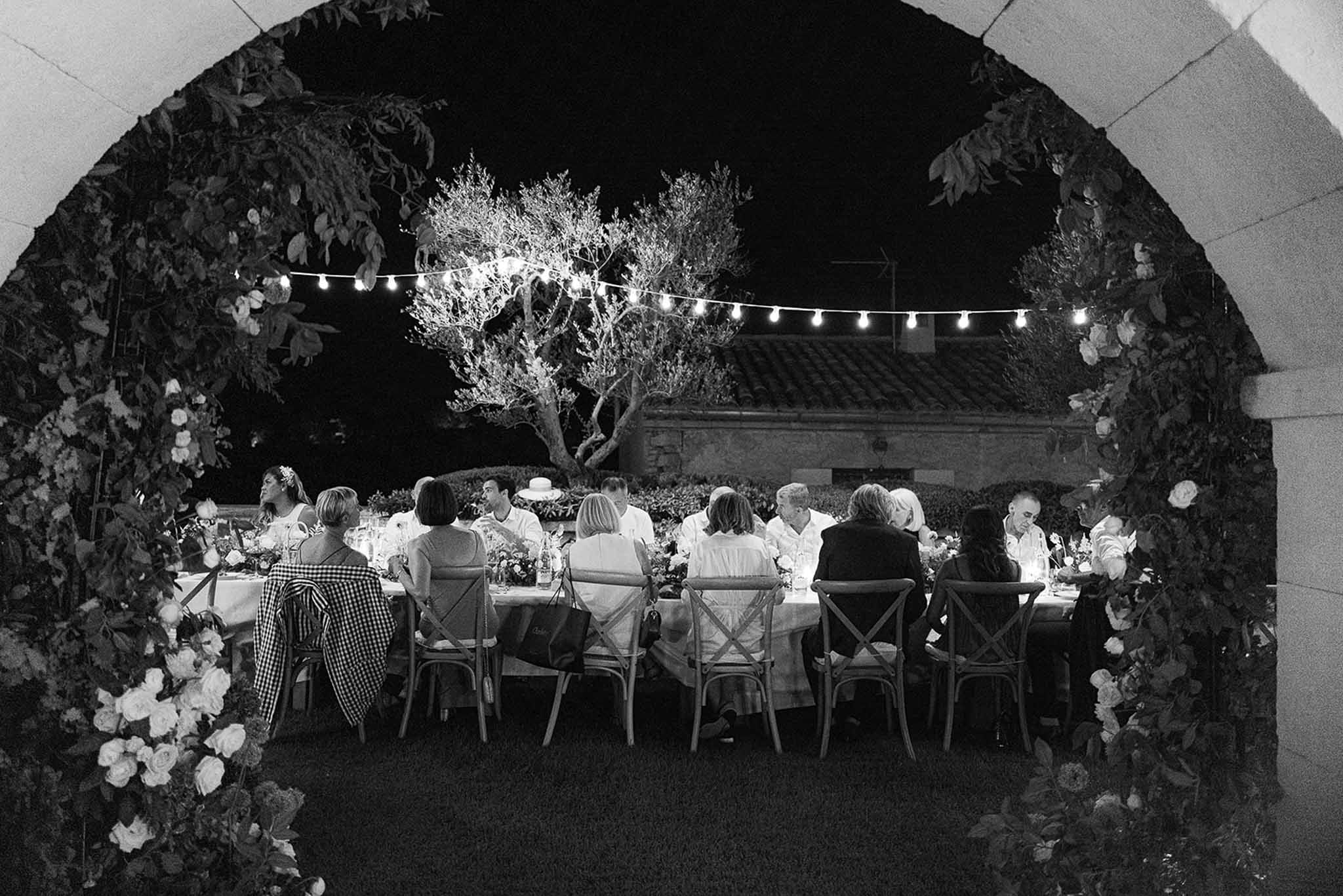 Reception dinner in outdoor stone courtyard with guests at long banquet table under archway with string lights
