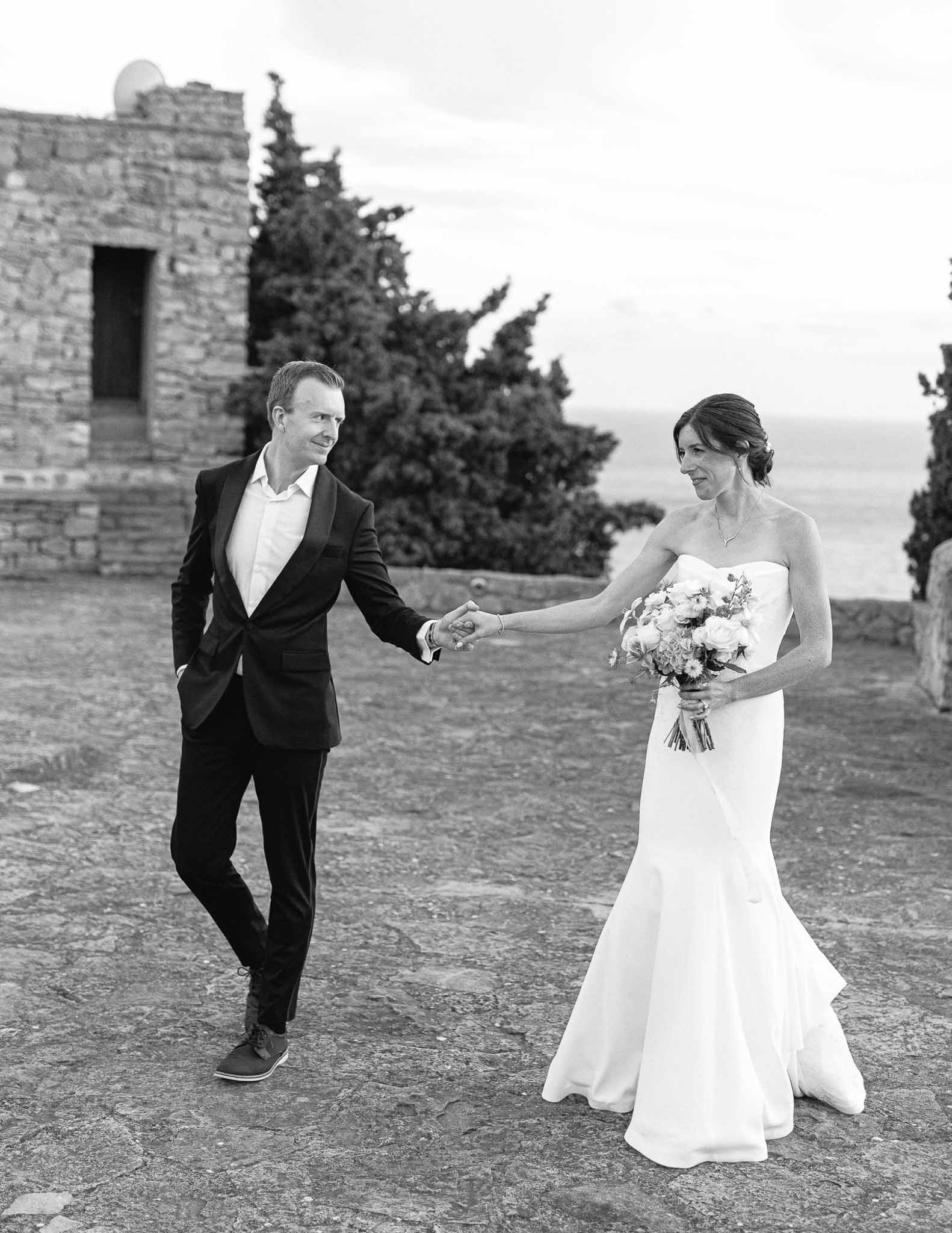 Bride and groom walking hand-in-hand outdoors at historic stone venue with cypress trees and water view