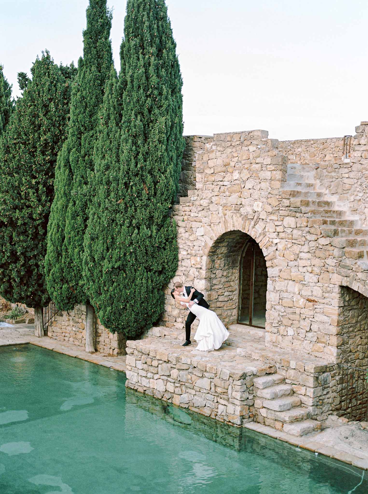 Groom kissing bride beside turquoise pool at Mediterranean stone venue with cypress trees