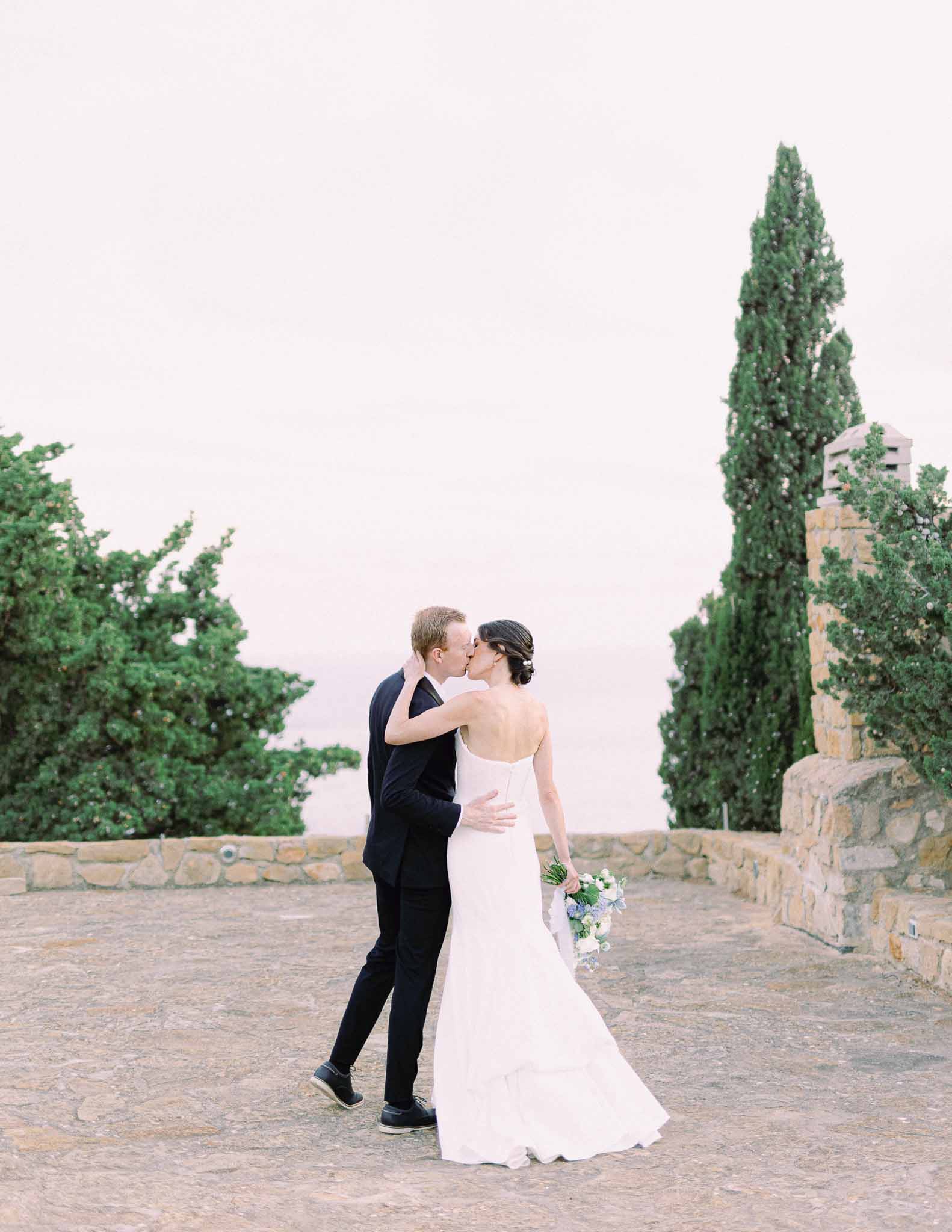 Bride and groom kissing on Mediterranean stone terrace with cypress trees and historic architecture