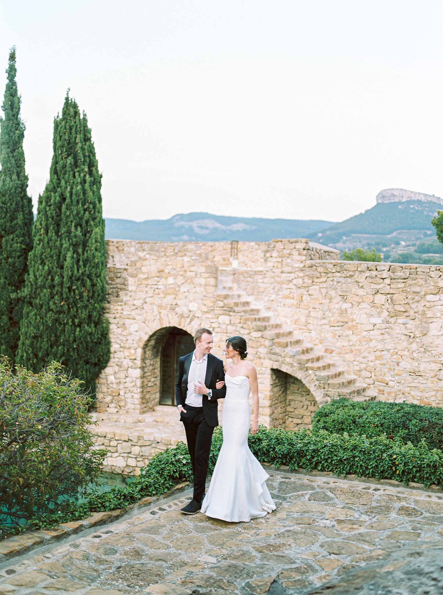 Bride and groom walking along stone pathway at Mediterranean countryside venue with ancient fortification