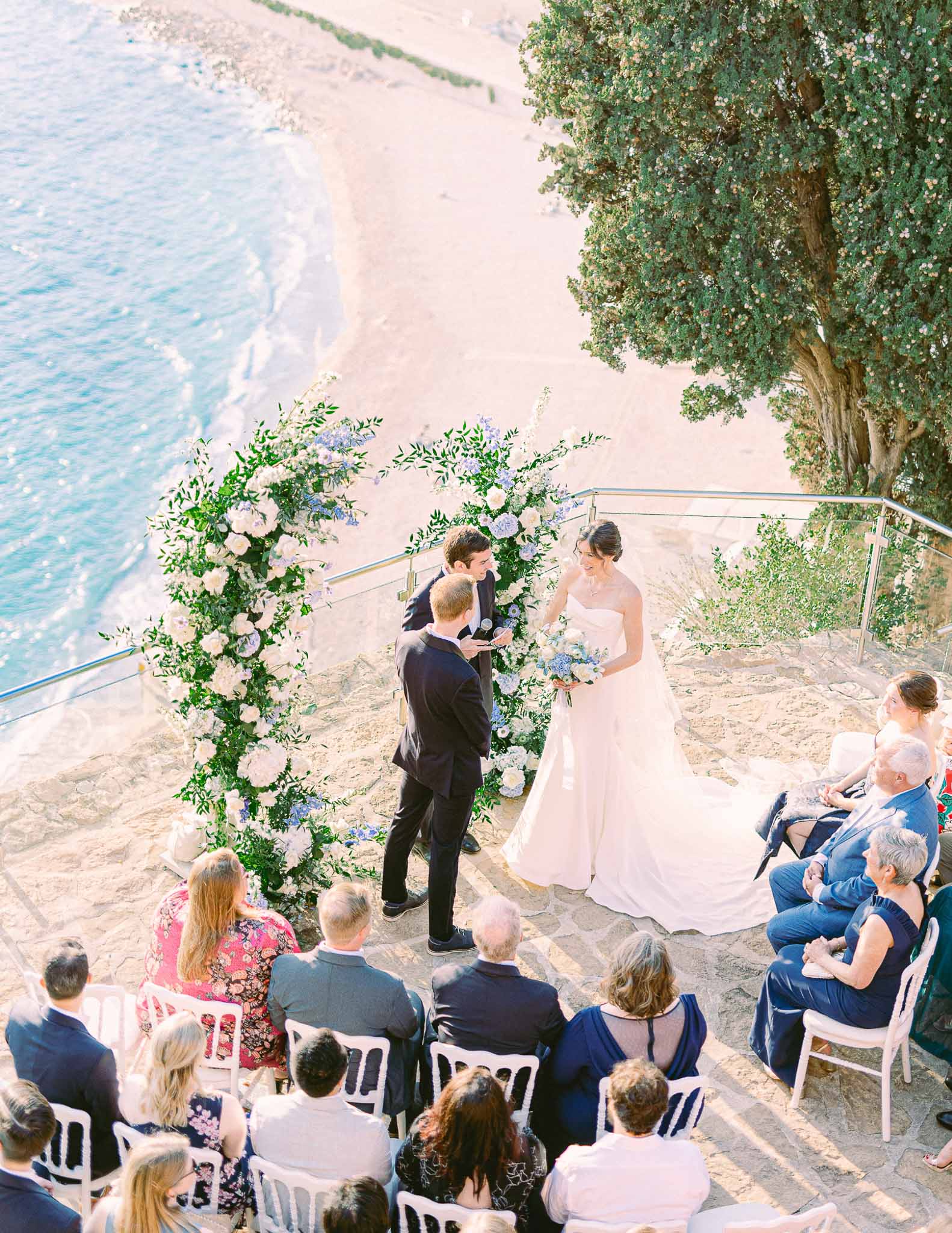 Aerial view of beachside wedding ceremony with floral arch and guests in white chairs on sand