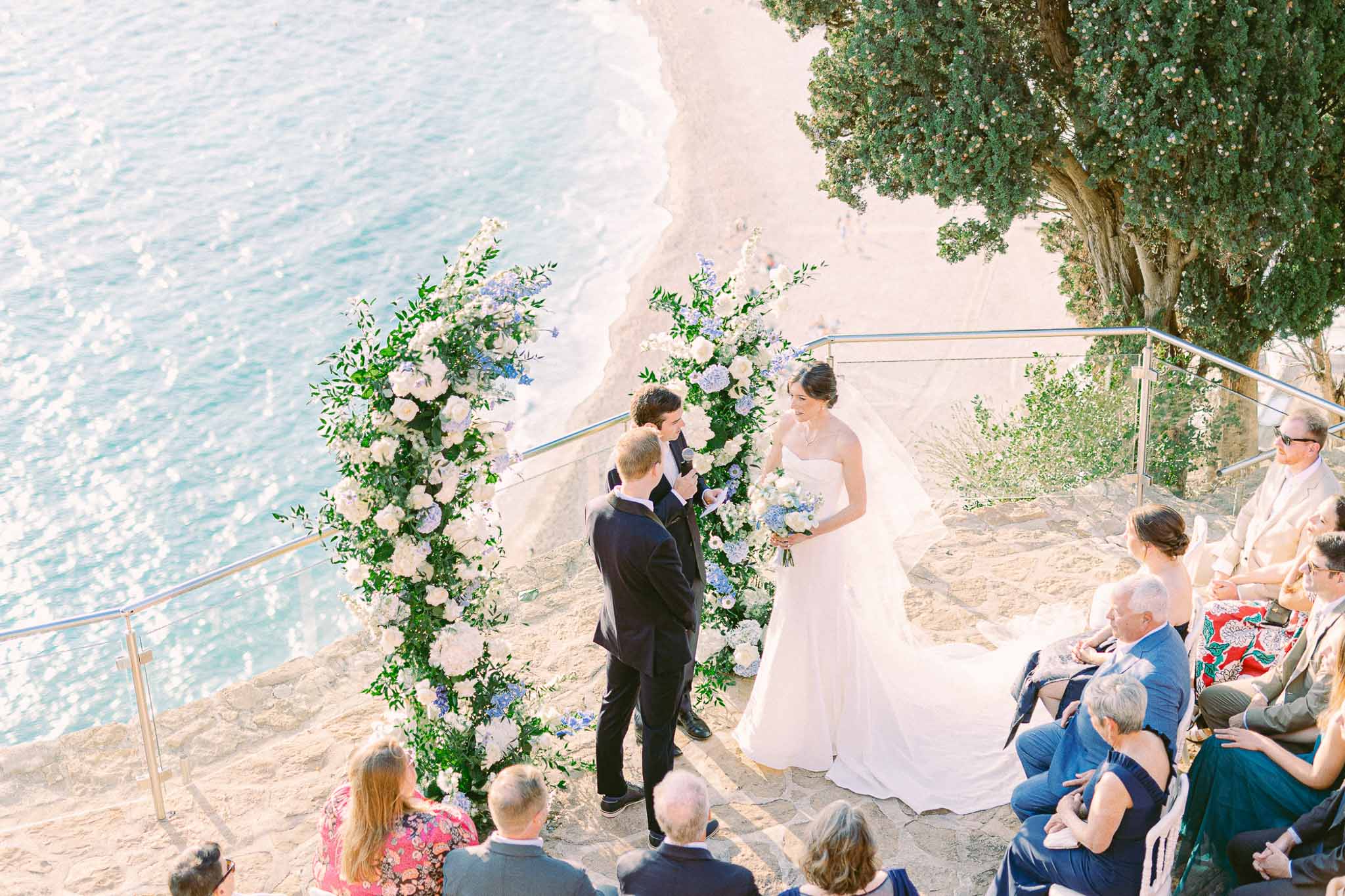 Bride and groom exchanging vows at seaside ceremony with floral arch and seated guests on beach